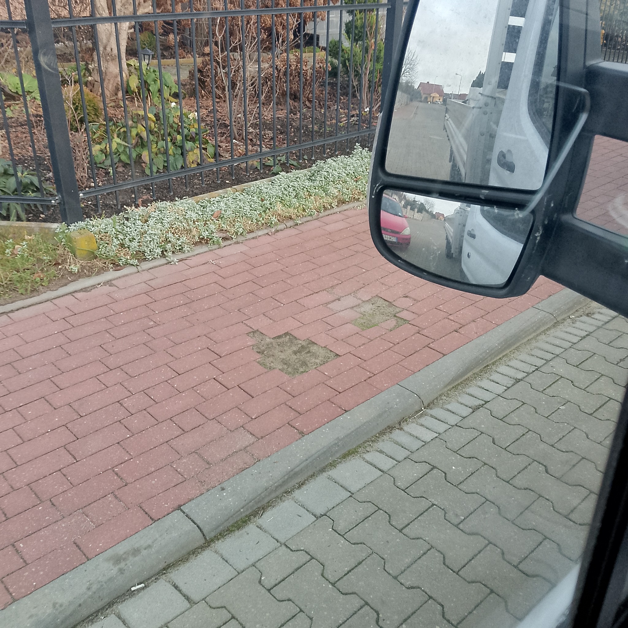 Czerwona kostka brukowa z ubytkami, widoczna z perspektywy lusterka samochodu. W tle czarne metalowe ogrodzenie i roślinność.
