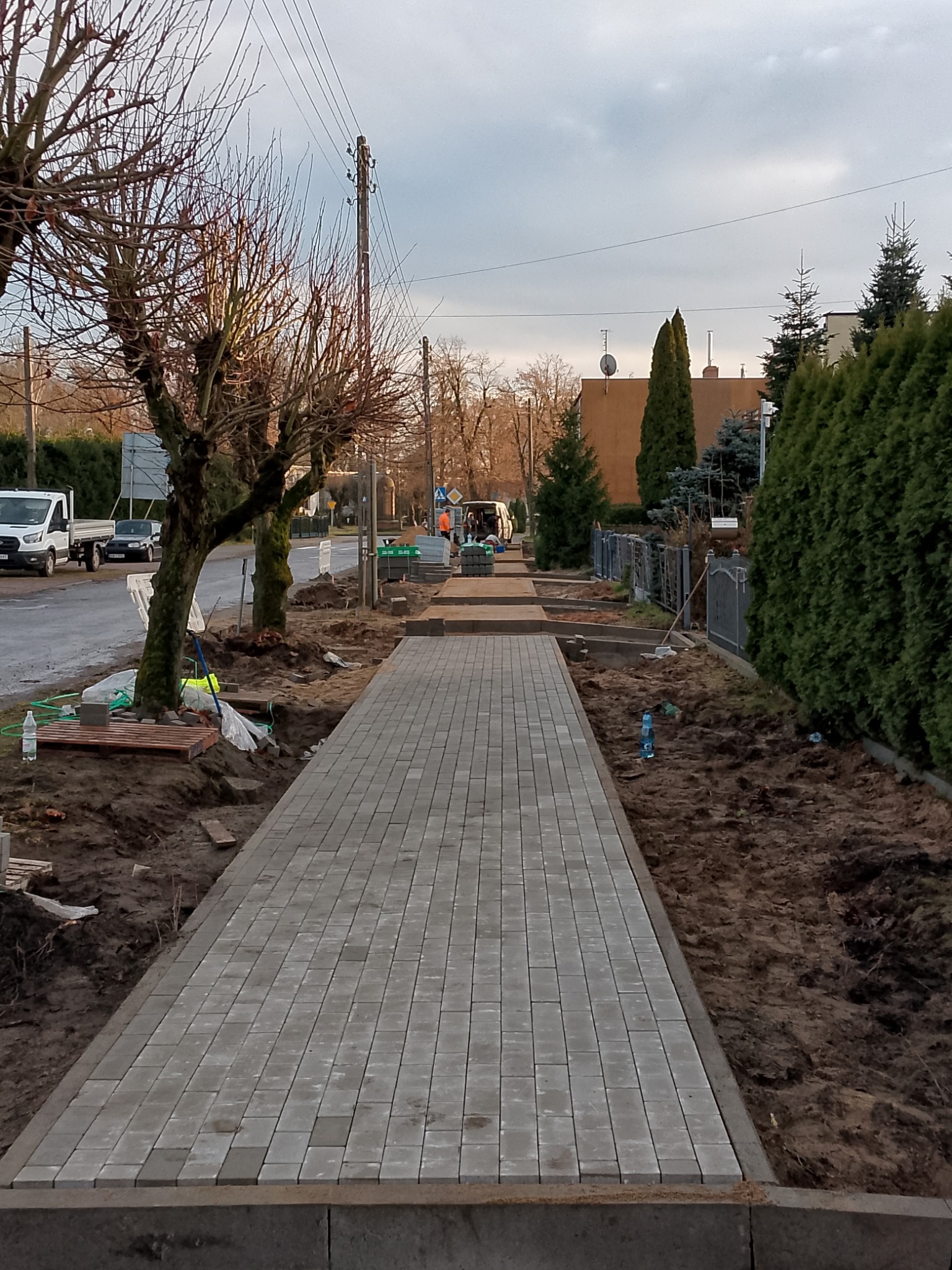 Świeżo ułożony chodnik z szarej kostki brukowej wzdłuż ulicy, z widocznymi elementami przygotowania terenu i pracownikami w oddali. Uwagę zwraca regularny wzór kostki.