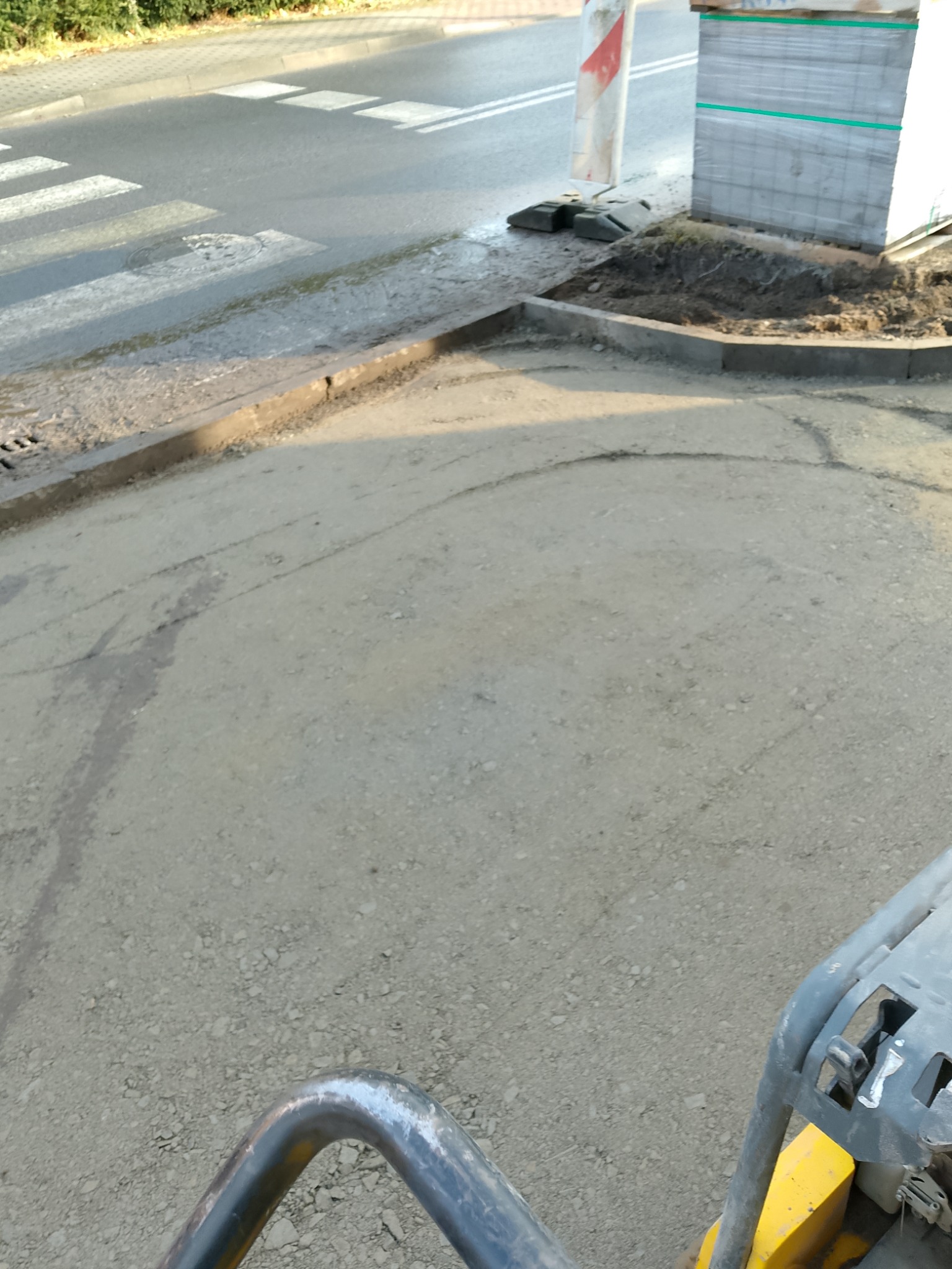 Przygotowany podkład pod kostkę brukową na chodniku przy przejściu dla pieszych, obok paleta z kostką i maszyna do ubijania.