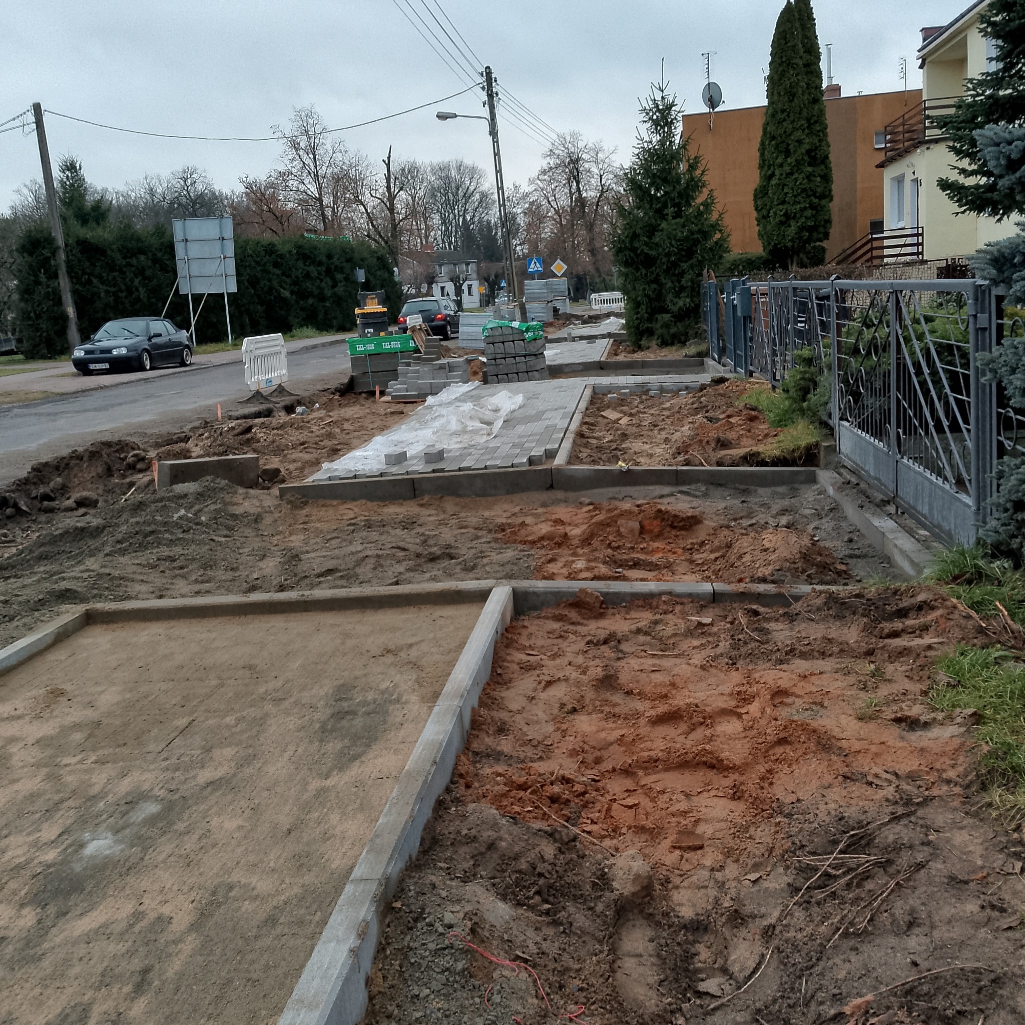 Układanie szarej kostki brukowej na posesji. Częściowo ułożona kostka, obrzeża z betonu, przygotowany teren. Widoczne narzędzia i materiały budowlane.