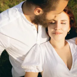 Z góry: Uśmiechnięta ruda kobieta w białej sukience leży na trawie, wtulona w brodatego mężczyznę w białej koszuli.