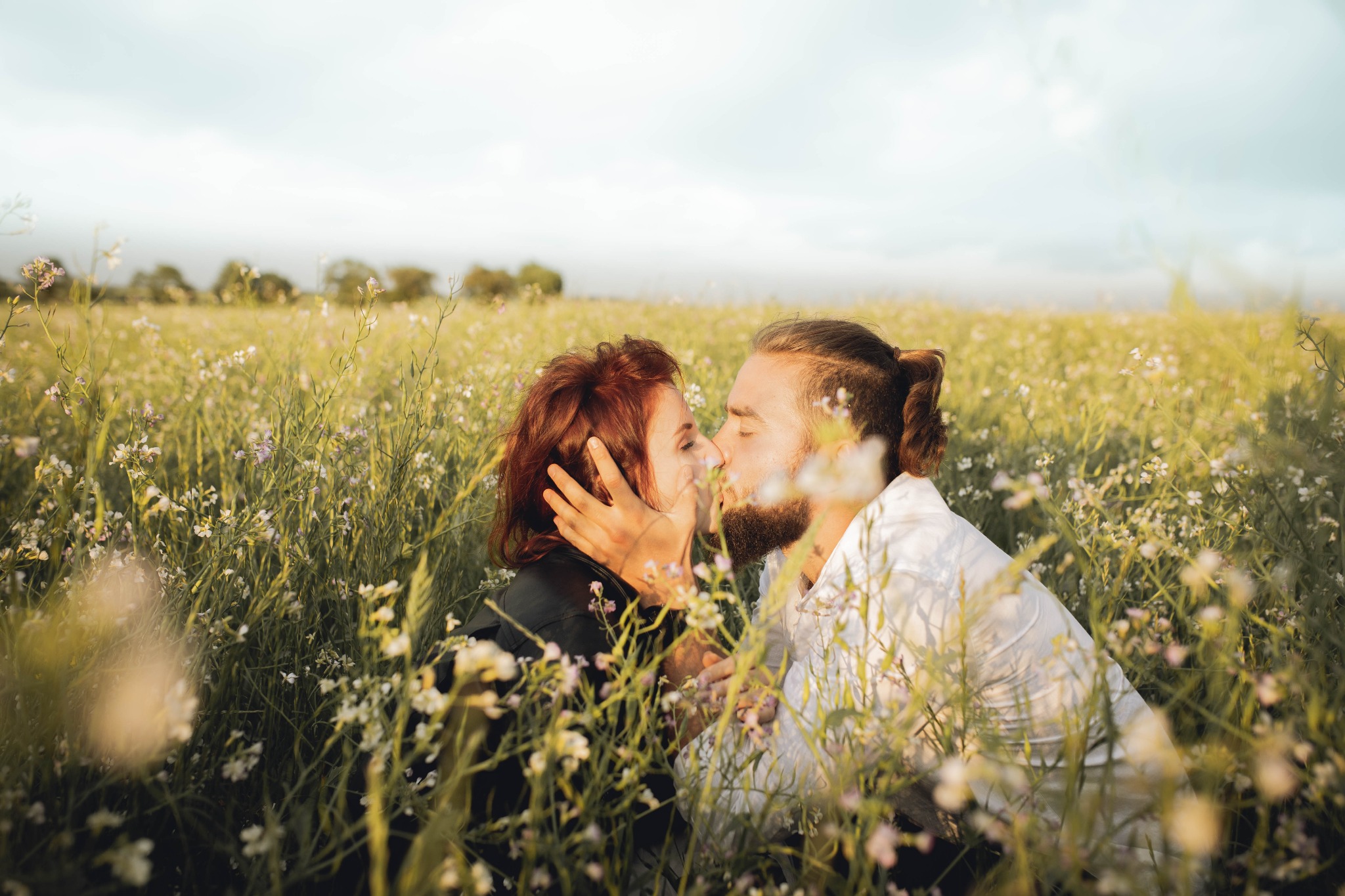 Zakochana para całuje się na kwitnącej łące, mężczyzna w białej koszuli, kobieta w czarnej kurtce, delikatne oświetlenie.