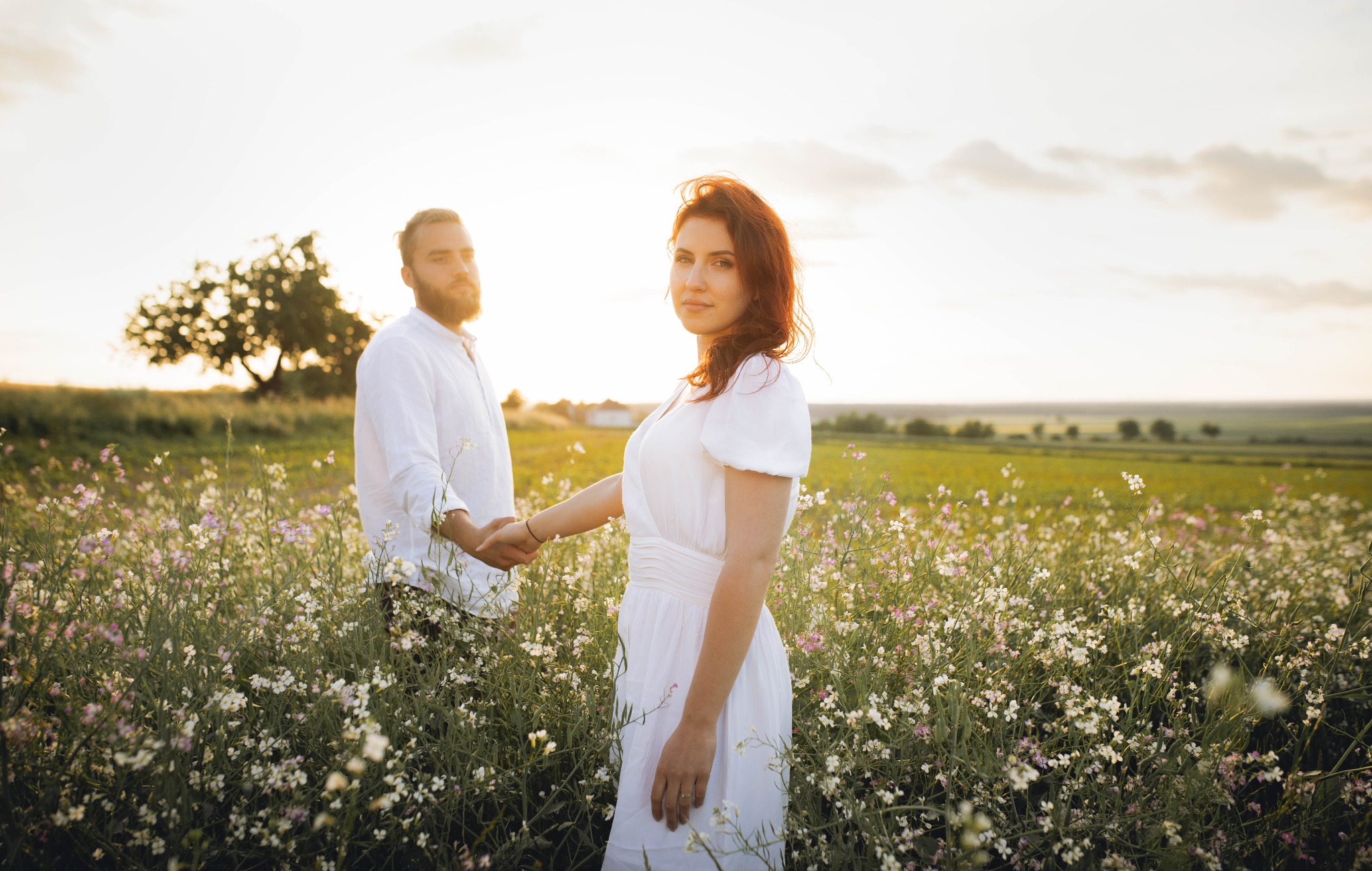 Para trzymająca się za ręce na kwitnącej łące w ciepłym, zachodzącym słońcu. Kobieta w białej sukience spogląda w obiektyw, mężczyzna w białej koszuli stoi w tle. W oddali widoczne drzewa...