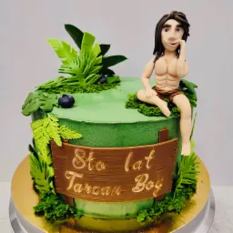 Tort urodzinowy z zielonym kremem, dekoracjami z liści i figurką Tarzana siedzącego na górze, z napisem 'Sto lat Tarzan Boy' na drewnianej tabliczce.
