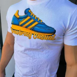 Mężczyzna w białej koszulce z nadrukiem stylizowanego buta sportowego w kolorach niebieskim i żółtym oraz napisem 'Tarnobrzeg Casual'.