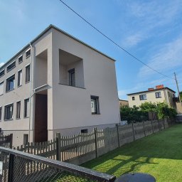 Bud-rem - Nowoczesny, dwukondygnacyjny dom z jasną elewacją i płaskim dachem, widoczny z perspektywy ogrodu, z betonowym ogrodzeniem w pierwszym planie.