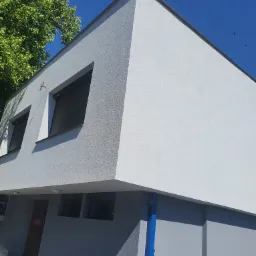 Nowoczesny budynek z szarą elewacją i ciemnymi roletami okiennymi, widziany pod kątem od dołu, z zielonym drzewem i błękitnym niebem w tle.