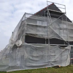 Dom w trakcie ocieplania elewacji, rusztowanie z siatką ochronną, widoczna czarna elewacja i fragment dachu z dachówką, teren trawiasty.