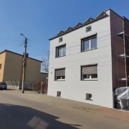 Dwupiętrowy dom po częściowym malowaniu elewacji na szaro, z brązowym fragmentem na bocznej ścianie, widoczne rusztowania i pracownik w trakcie malowania, na dachu panele słoneczne, ulica...