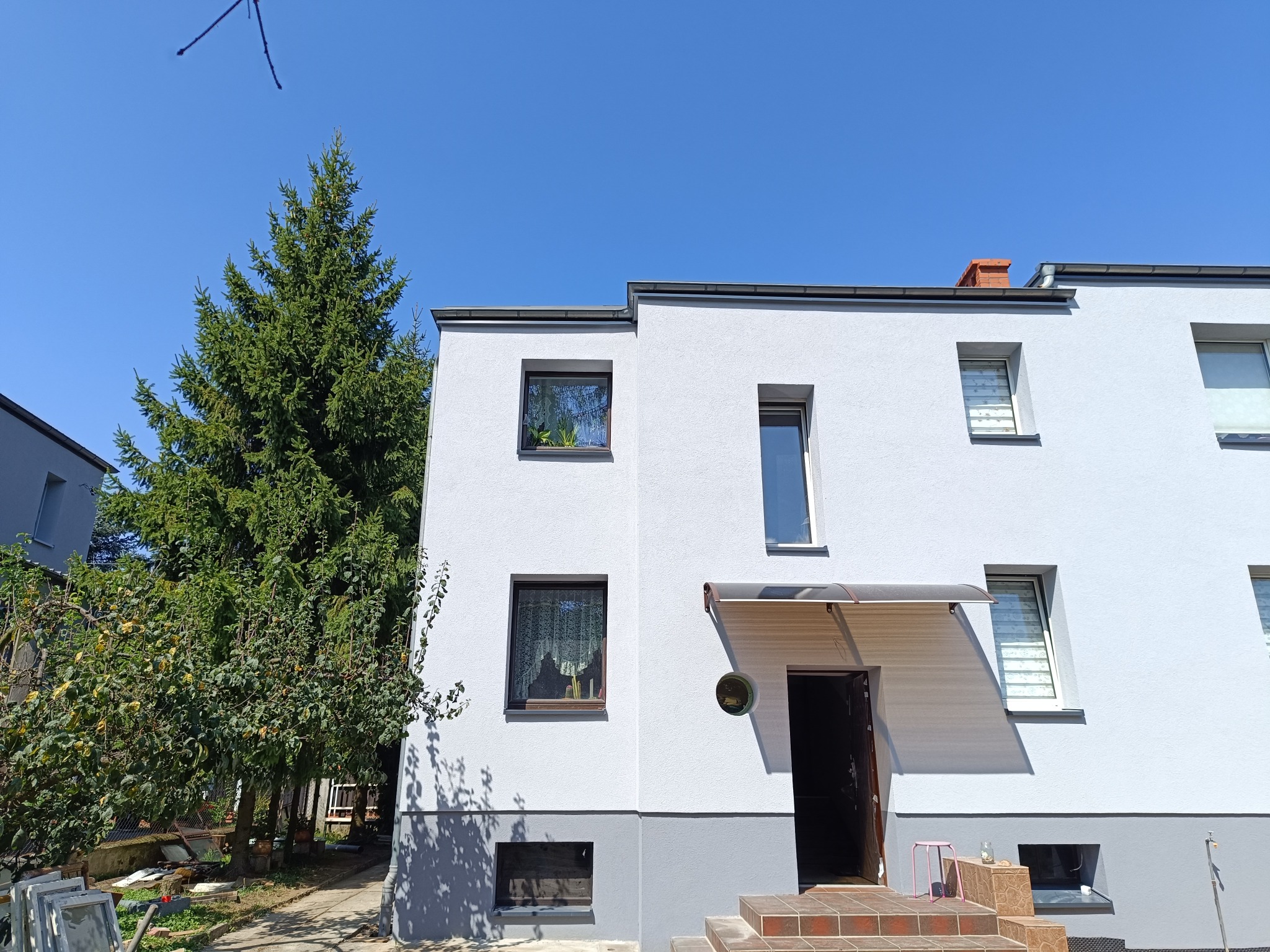 Nowoczesny, dwukondygnacyjny dom z jasną elewacją i ciemnymi ramami okiennymi, widoczny z poziomu gruntu, z zielonymi drzewami po lewej stronie i schodami wejściowymi wyłożonymi brązowymi płytkami.