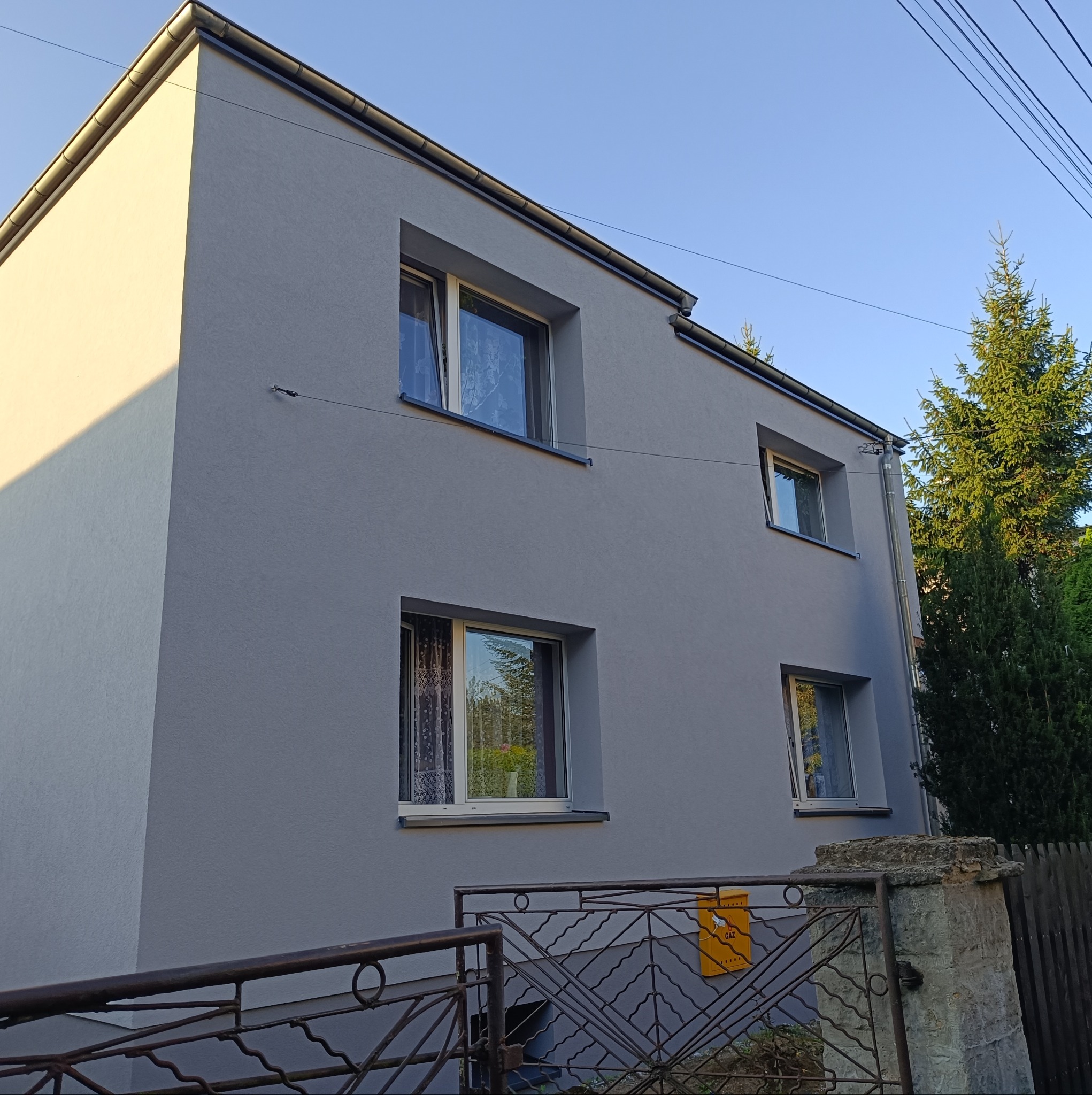 Dwupiętrowy dom z szarą, gładką elewacją, widok z dołu, z metalowym ogrodzeniem i żółtą skrzynką na listy przy bramie.