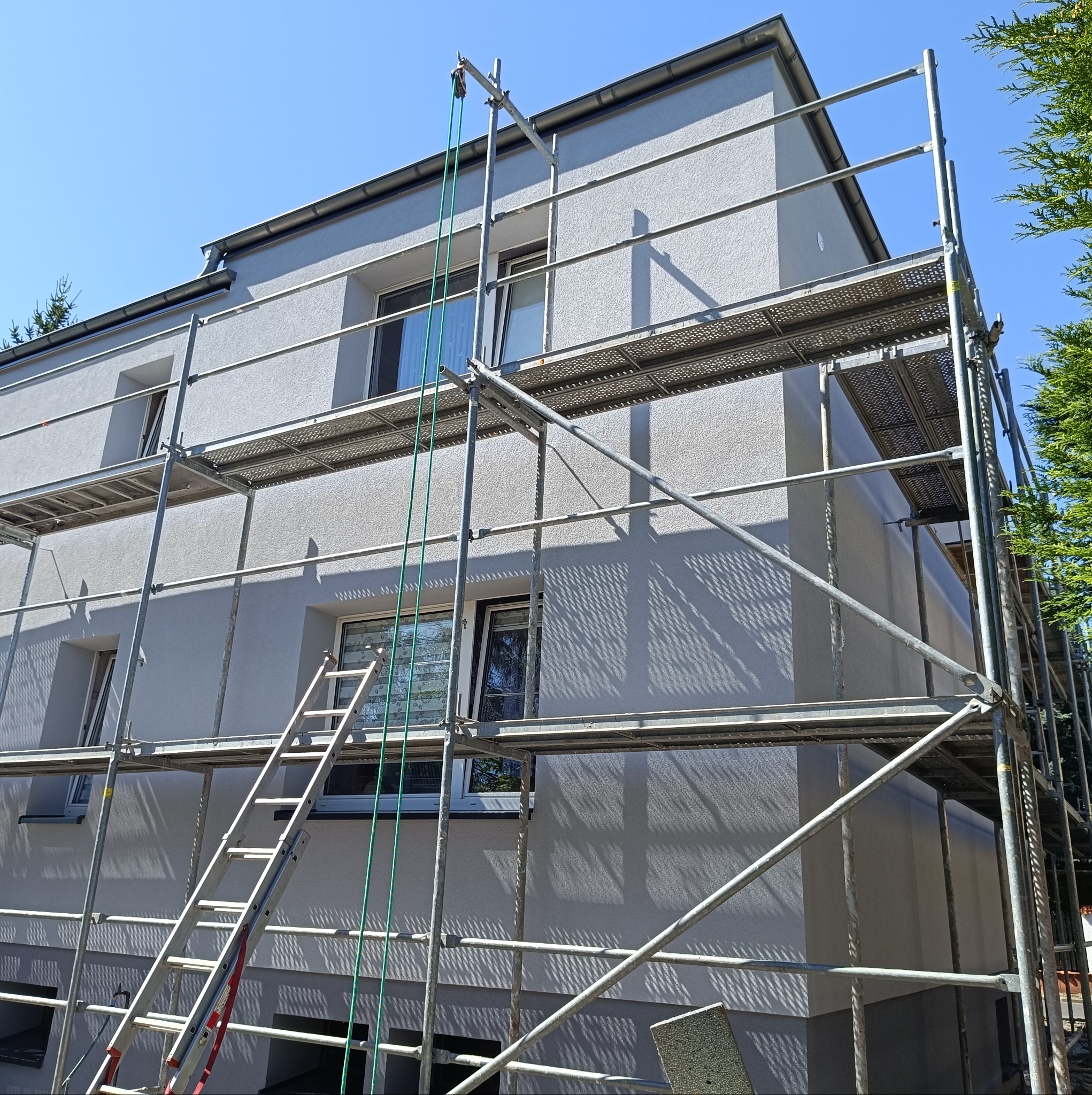 Dom jednorodzinny w trakcie prac elewacyjnych, z widocznymi rusztowaniami, drabiną i fragmentem ocieplenia, na tle błękitnego nieba i zieleni.