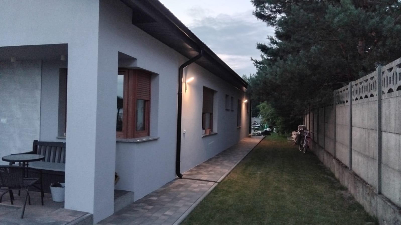 Nowoczesny dom jednorodzinny z szarą elewacją, drewnianymi okiennicami i oświetleniem zewnętrznym, widok z boku na tle zieleni i betonowego ogrodzenia.