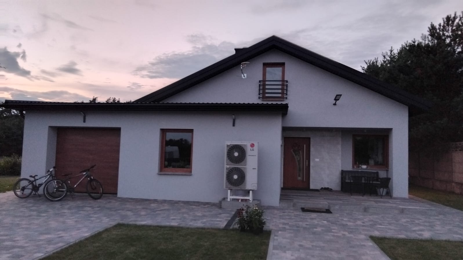 Nowoczesny dom jednorodzinny z szarą elewacją, brązowymi ramami okiennymi i drzwiami, oraz klimatyzatorem LG na zewnątrz. Na podjeździe stoją dwa rowery, a w tle widać drzewa i zachodzące słońce.
