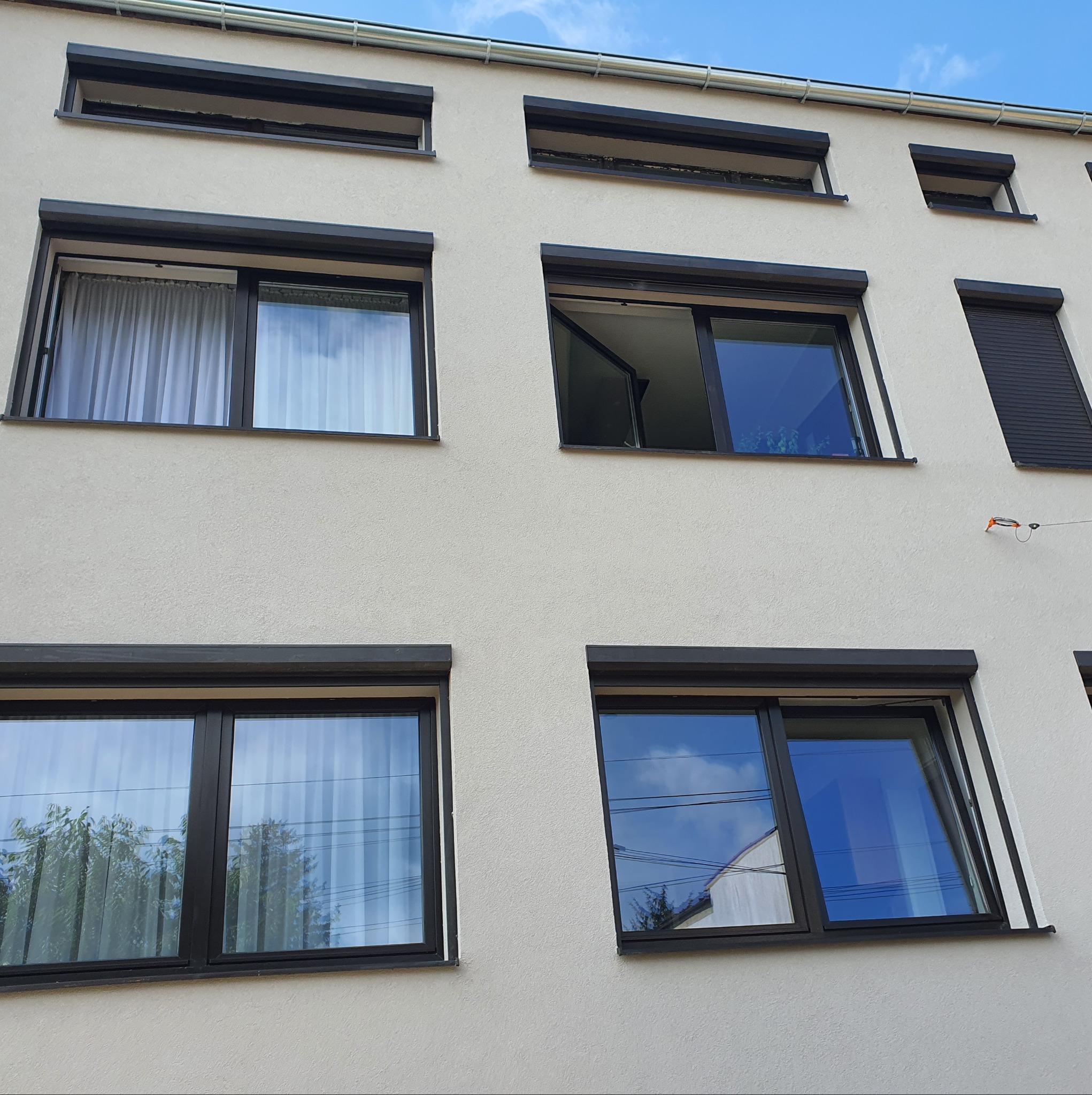 Elewacja budynku z oknami z czarnymi ramami i częściowo opuszczonymi roletami zewnętrznymi. Widoczne odbicia nieba i drzew w szybach.