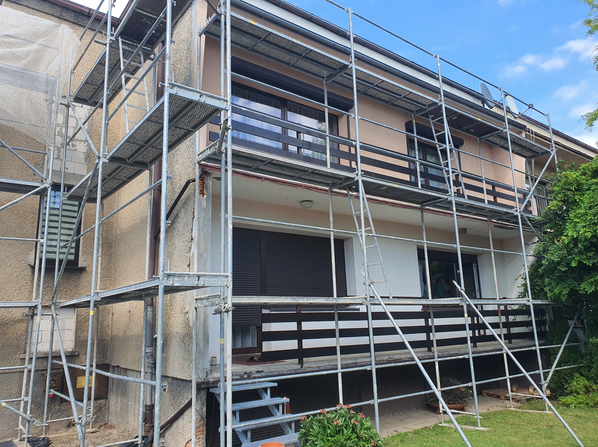 Dwupiętrowy dom w trakcie remontu elewacji, otoczony metalowymi rusztowaniami, z widocznym fragmentem elewacji przed i po renowacji.