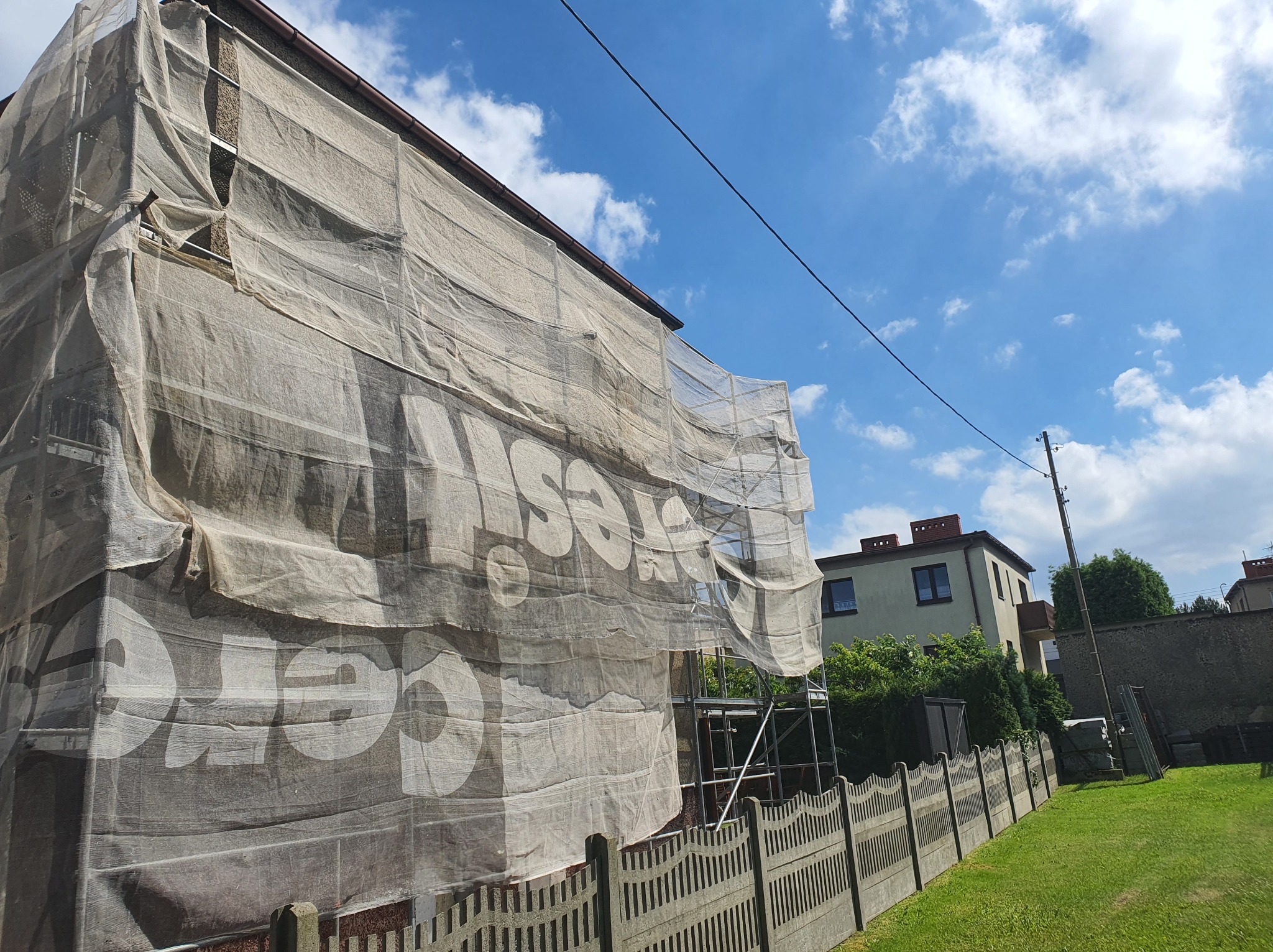 Budynek mieszkalny w trakcie prac elewacyjnych, osłonięty siatką z nadrukiem reklamowym, widoczne rusztowania, fragment ogrodzenia i sąsiedni budynek w tle, słoneczny dzień.