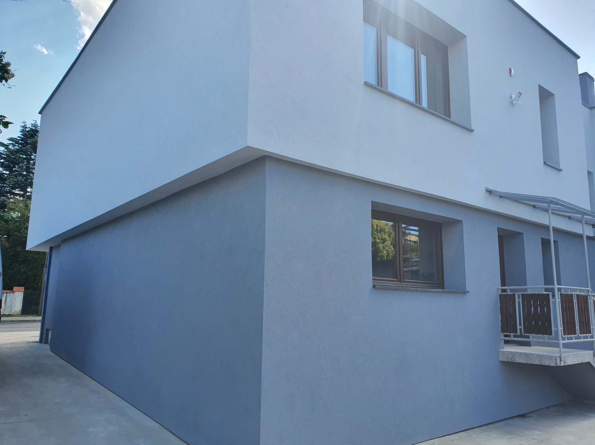 Dwukondygnacyjny budynek z elewacją w dwóch odcieniach szarości, z widocznymi oknami z brązowymi ramami i niewielkim balkonem z metalową balustradą.