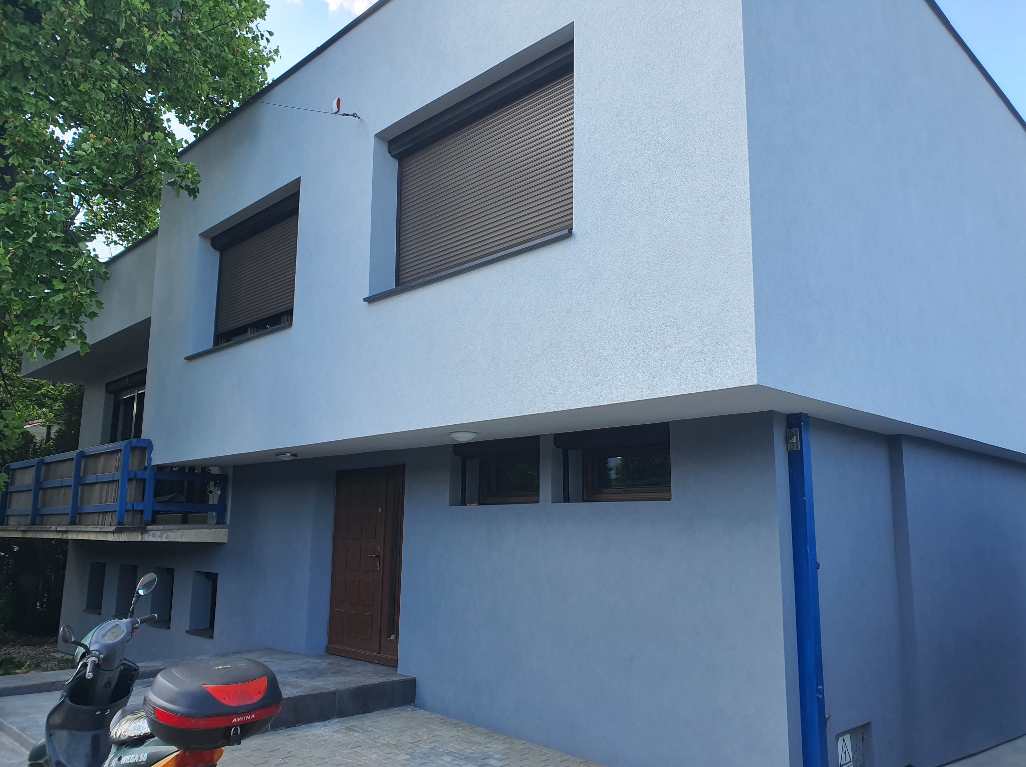 Dwukondygnacyjny budynek z szarą elewacją, brązowymi roletami i drzwiami wejściowymi, na pierwszym planie skuter