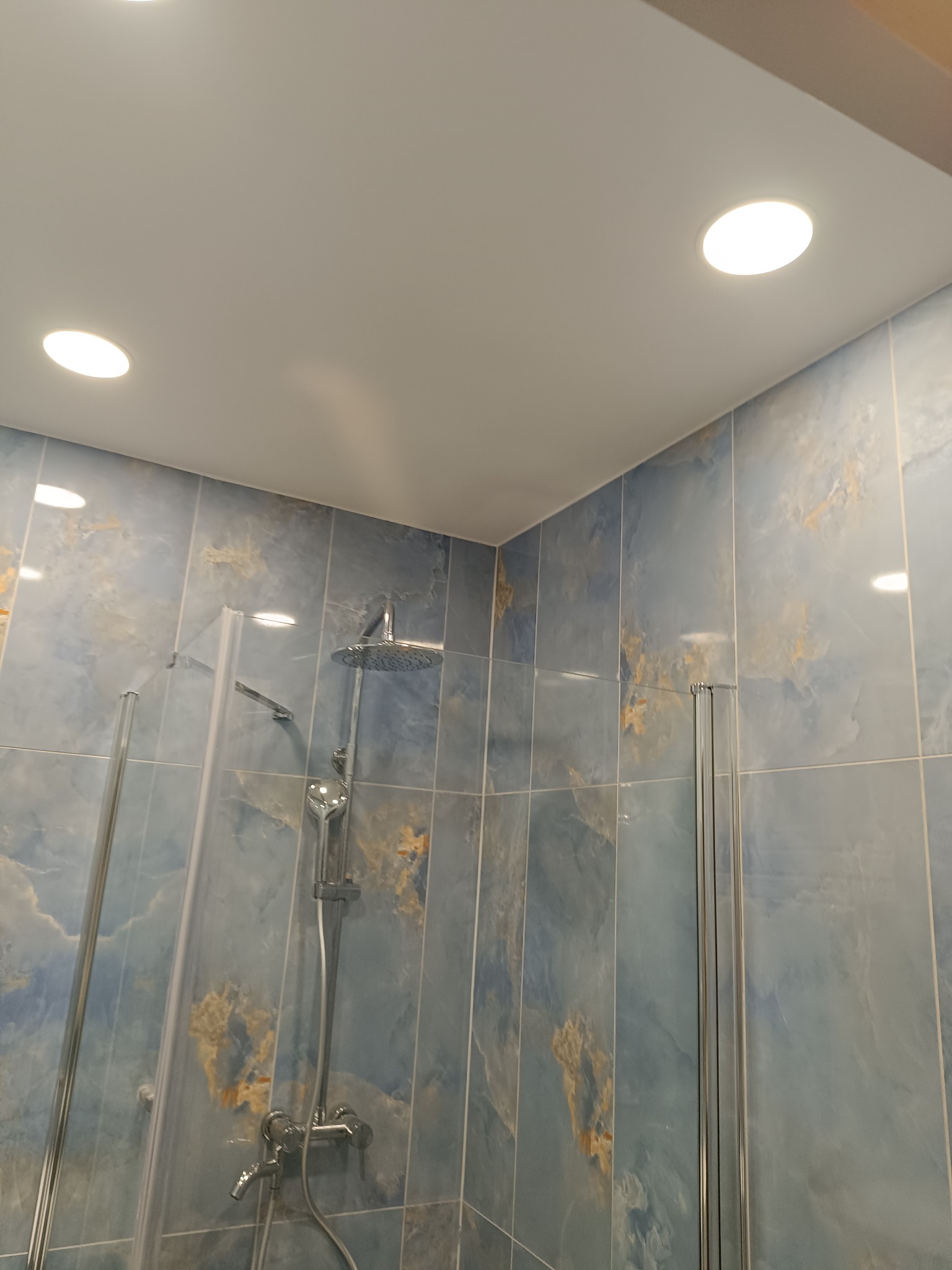 Błękitna łazienka z kabiną prysznicową i deszczownicą. Płytki imitujące marmur, punktowe oświetlenie sufitowe. Nowoczesny design.