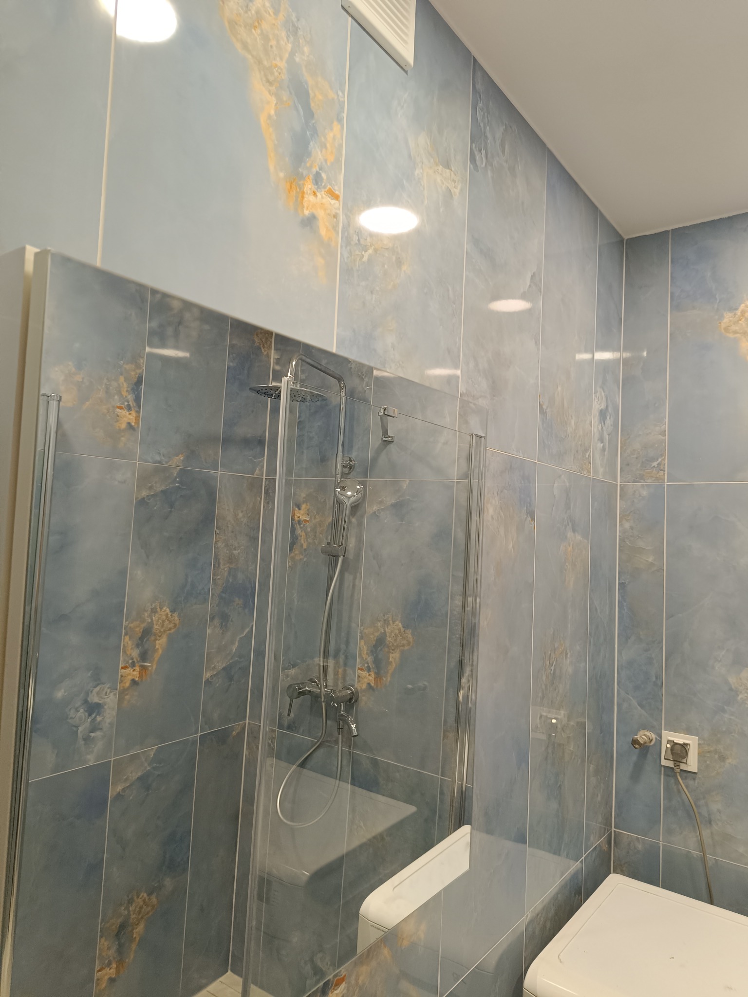 Wykończona łazienka z kabiną prysznicową i płytkami imitującymi marmur w odcieniach błękitu i złota. Widoczna bateria prysznicowa i fragment pralki.