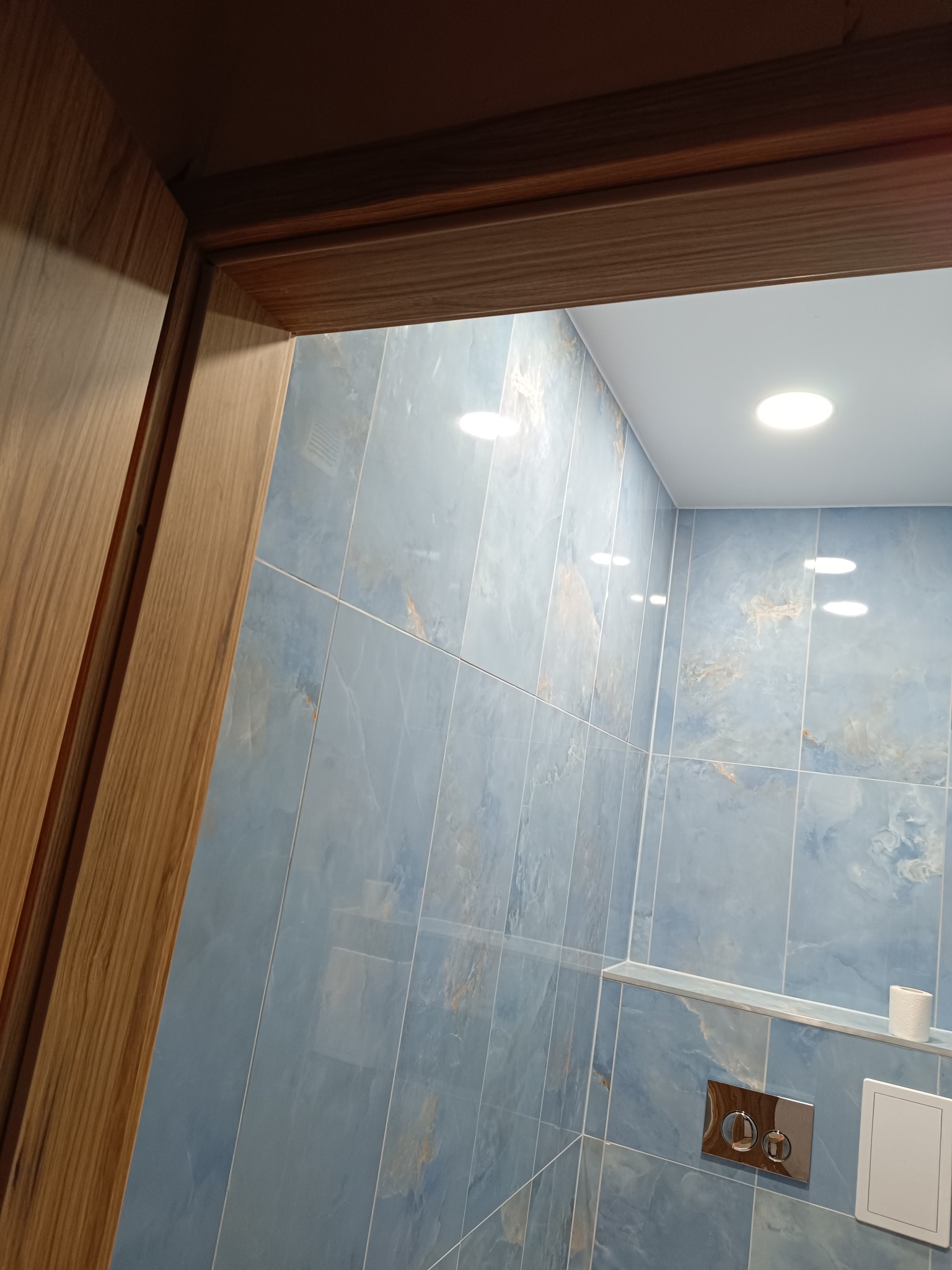 Błękitne płytki imitujące marmur w wykończonej łazience, widoczne przez drewnianą futrynę. Na ścianie podwieszana toaleta i półka z akcesoriami. Oświetlenie sufitowe.