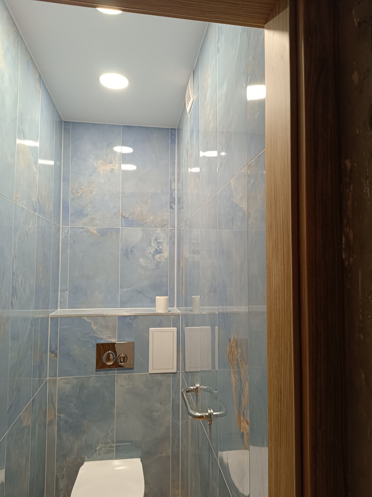 Niebieska łazienka z marmurowymi płytkami, podwieszaną toaletą i chromowanymi detalami, widziana zza drewnianej futryny. Minimalistyczny design z oświetleniem punktowym.