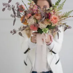 Kobieta w białej marynarce i jeansach zasłania twarz bukietem z różowych, brzoskwiniowych i białych kwiatów z zielonymi dodatkami i delikatną różową wstążką.