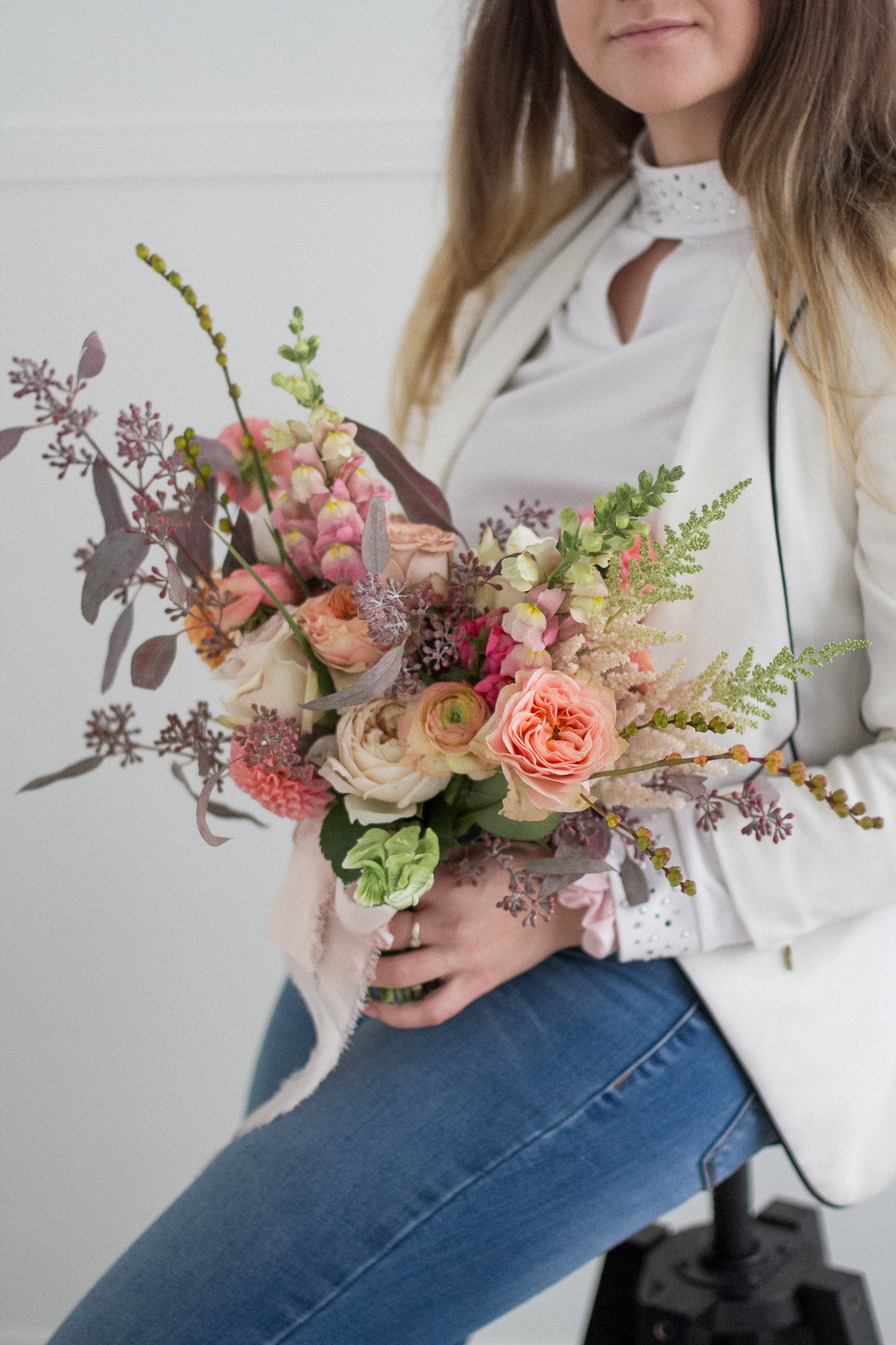 Kobieta w białej marynarce i jeansach trzyma w dłoniach pastelowy bukiet z róż, lwich paszczy i eukaliptusa, przewiązany jedwabną wstążką.