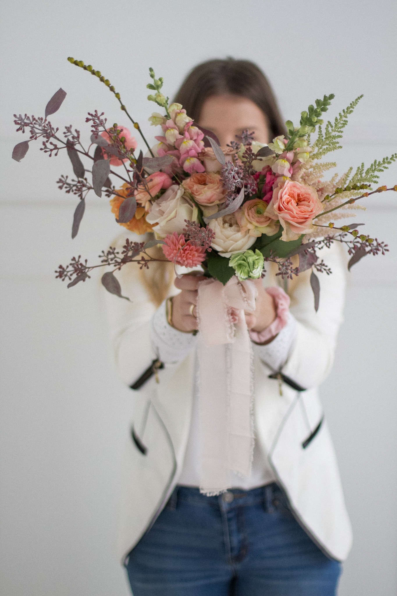 Kobieta w białej marynarce i jeansach zasłania twarz bukietem z różowych, brzoskwiniowych i białych kwiatów z zielonymi dodatkami i delikatną różową wstążką.