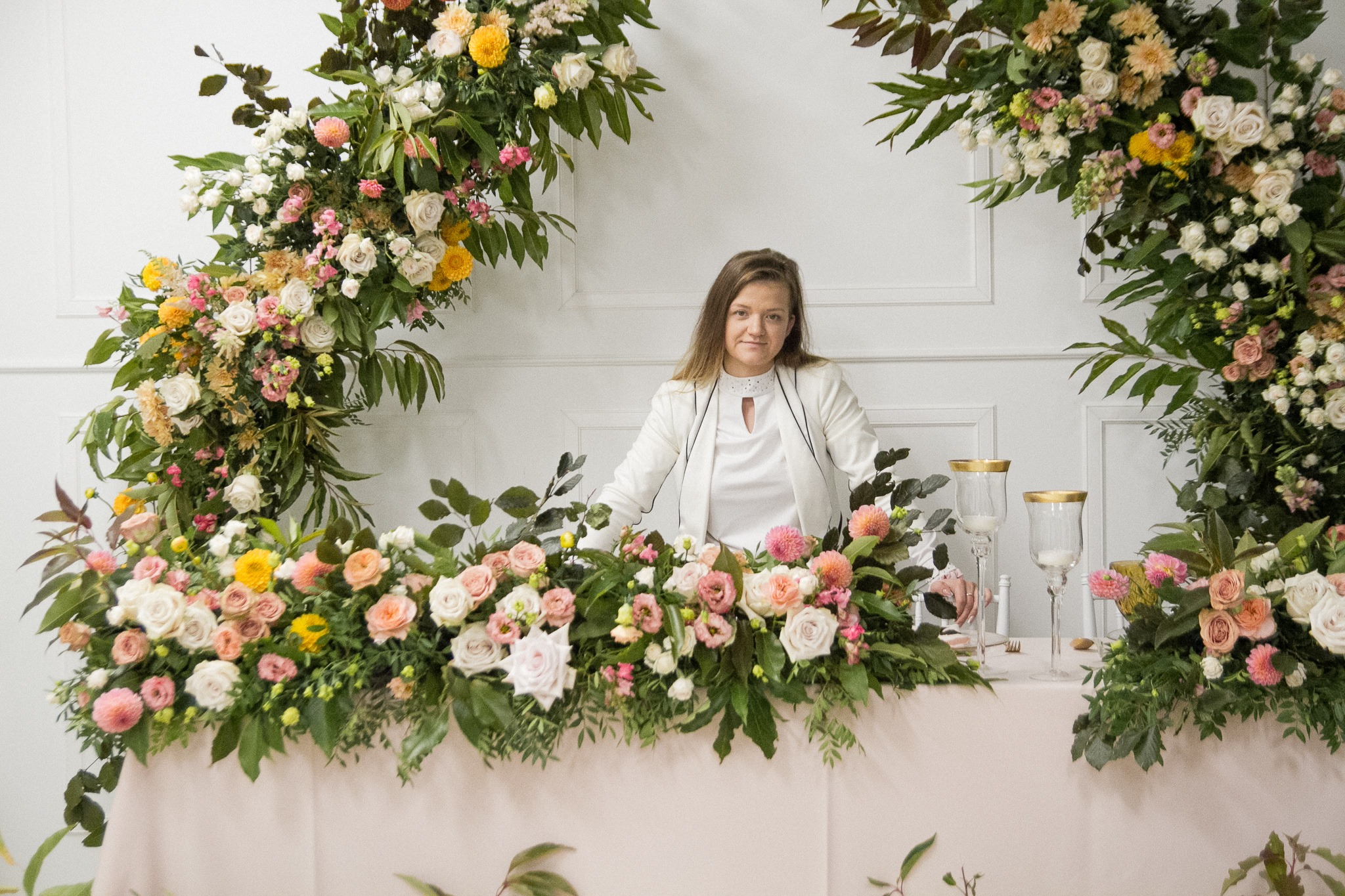 Kobieta w białym żakiecie stoi za stołem udekorowanym kompozycją z różowych, żółtych i białych kwiatów; nad stołem dwa łuki z podobnych kwiatów na tle białej ściany.