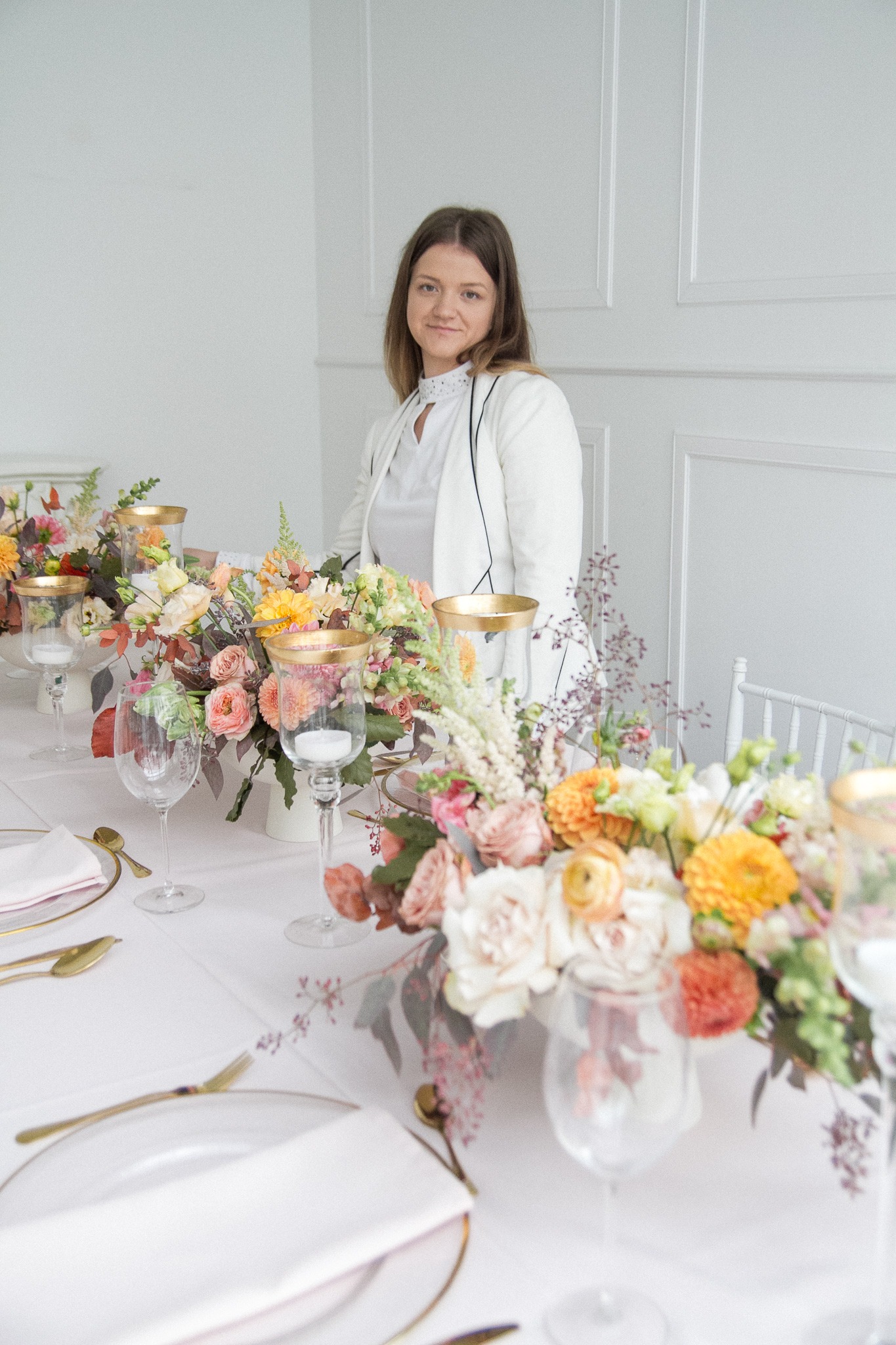 Uśmiechnięta kobieta w białej marynarce stoi za stołem udekorowanym kwiatowymi kompozycjami w odcieniach różu, pomarańczy i żółci, ze złotymi świecznikami i nakryciami, przygotowanym...