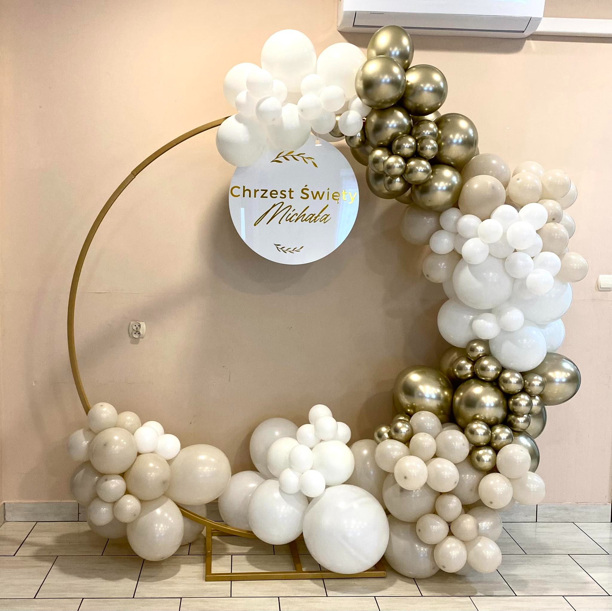 Dekoracja balonowa w kolorach bieli, beżu i złota na Chrzest Święty Michała, z okrągłym złotym stelażem i tabliczką z napisem, klimatyzator na ścianie w tle.