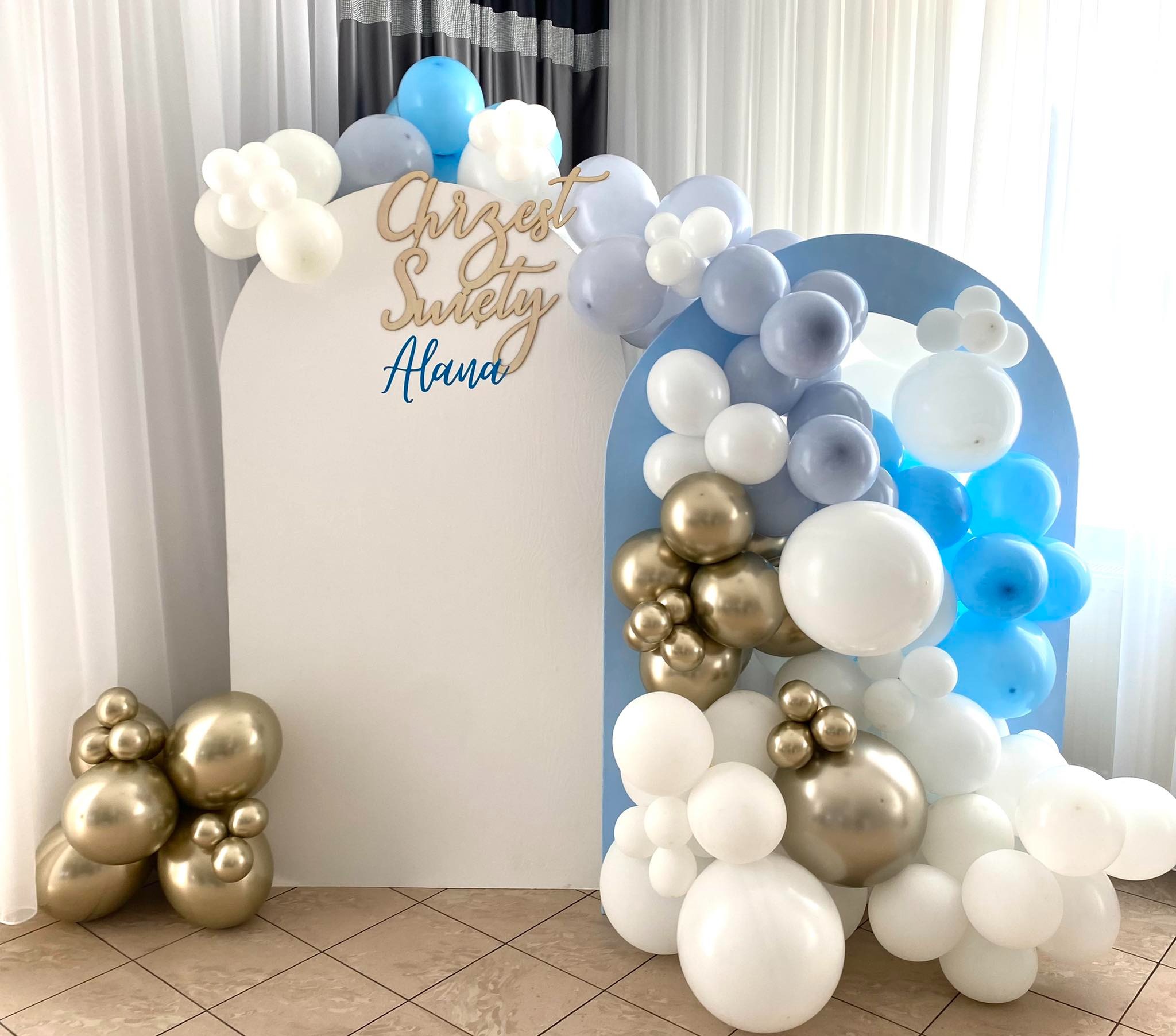 Dekoracja balonowa na chrzest z białymi, błękitnymi i złotymi balonami, drewniany napis 'Chrzest Święty Alana' na białym tle w kształcie łuku.