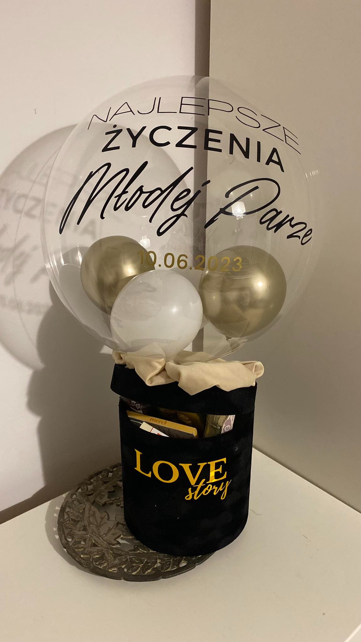 Dekoracja ślubna: przezroczysty balon z napisem 'Najlepsze Życzenia Młodej Parze' i datą 10.06.2023, zawierający trzy balony w kolorach złotym i białym, umieszczony w czarnym welurowym pudełku...