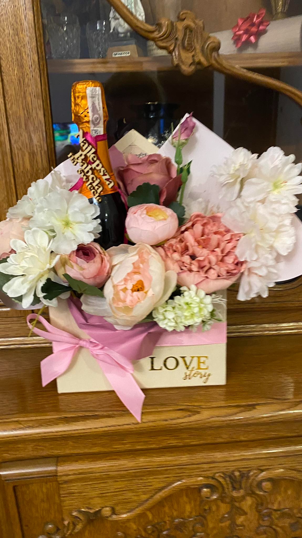 Kompozycja kwiatowa w pudełku z napisem 'LOVE story' i butelką szampana 'Najlepsza Babcia' na tle drewnianego mebla.