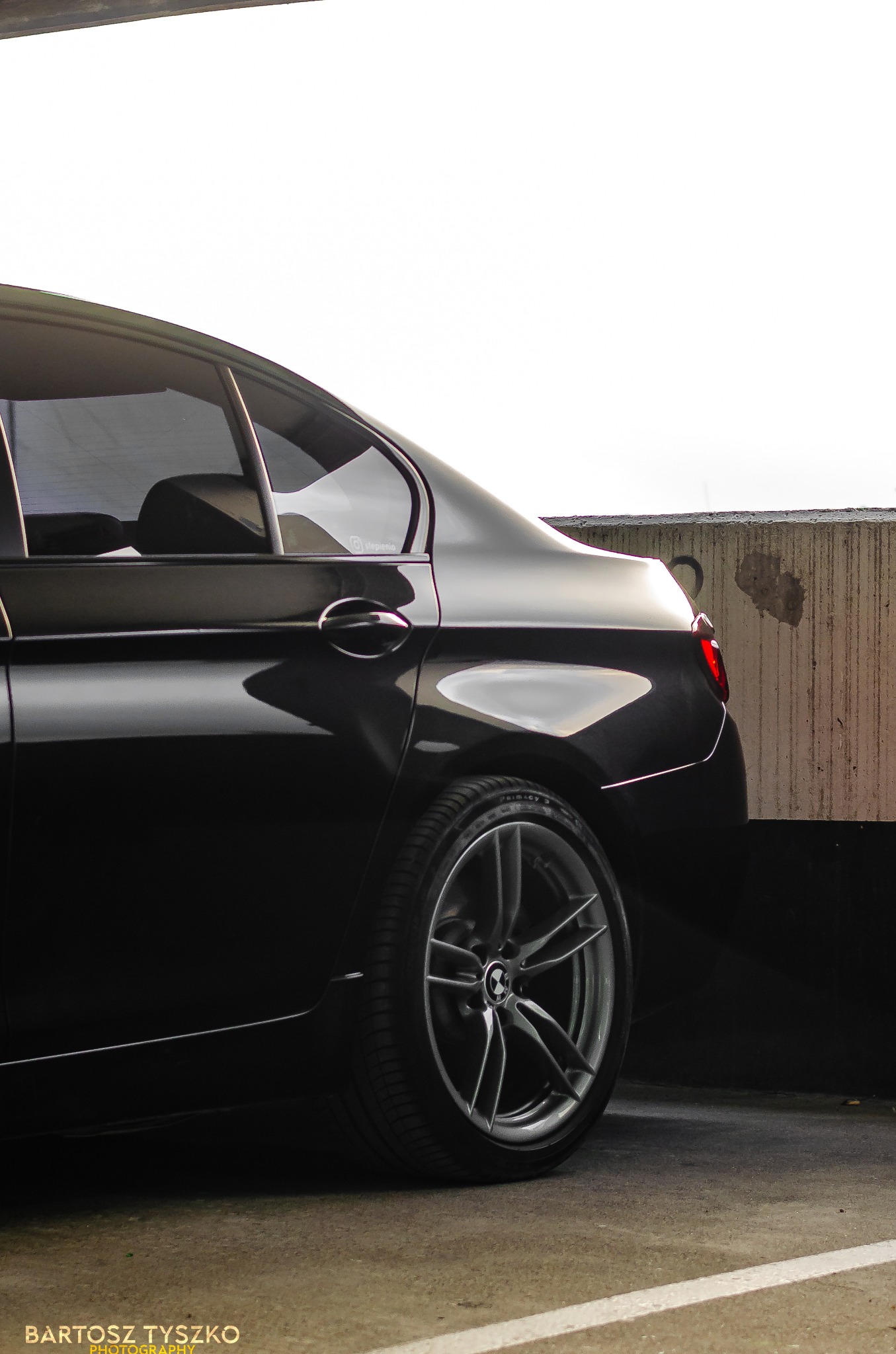 Częściowy widok czarnego samochodu BMW serii 5 z felgami aluminiowymi, zaparkowanego na betonowym parkingu z widocznymi śladami użytkowania, z logo fotografa w lewym dolnym rogu.
