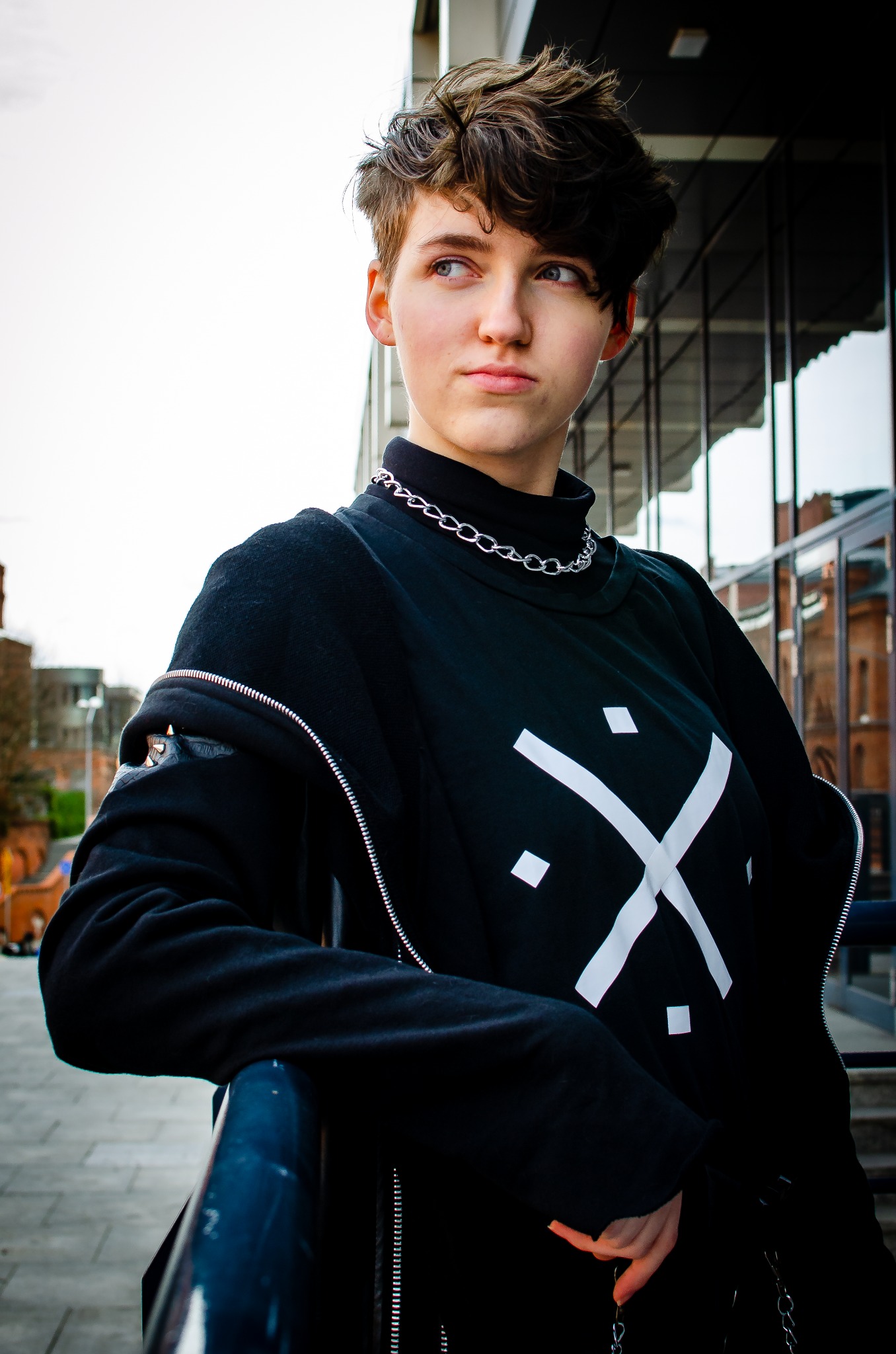 Portret osoby z krótkimi, ciemnymi włosami, ubranej w czarny golf i bluzę z białym geometrycznym wzorem oraz srebrny łańcuch, opierającej się o barierkę na tle miejskim.