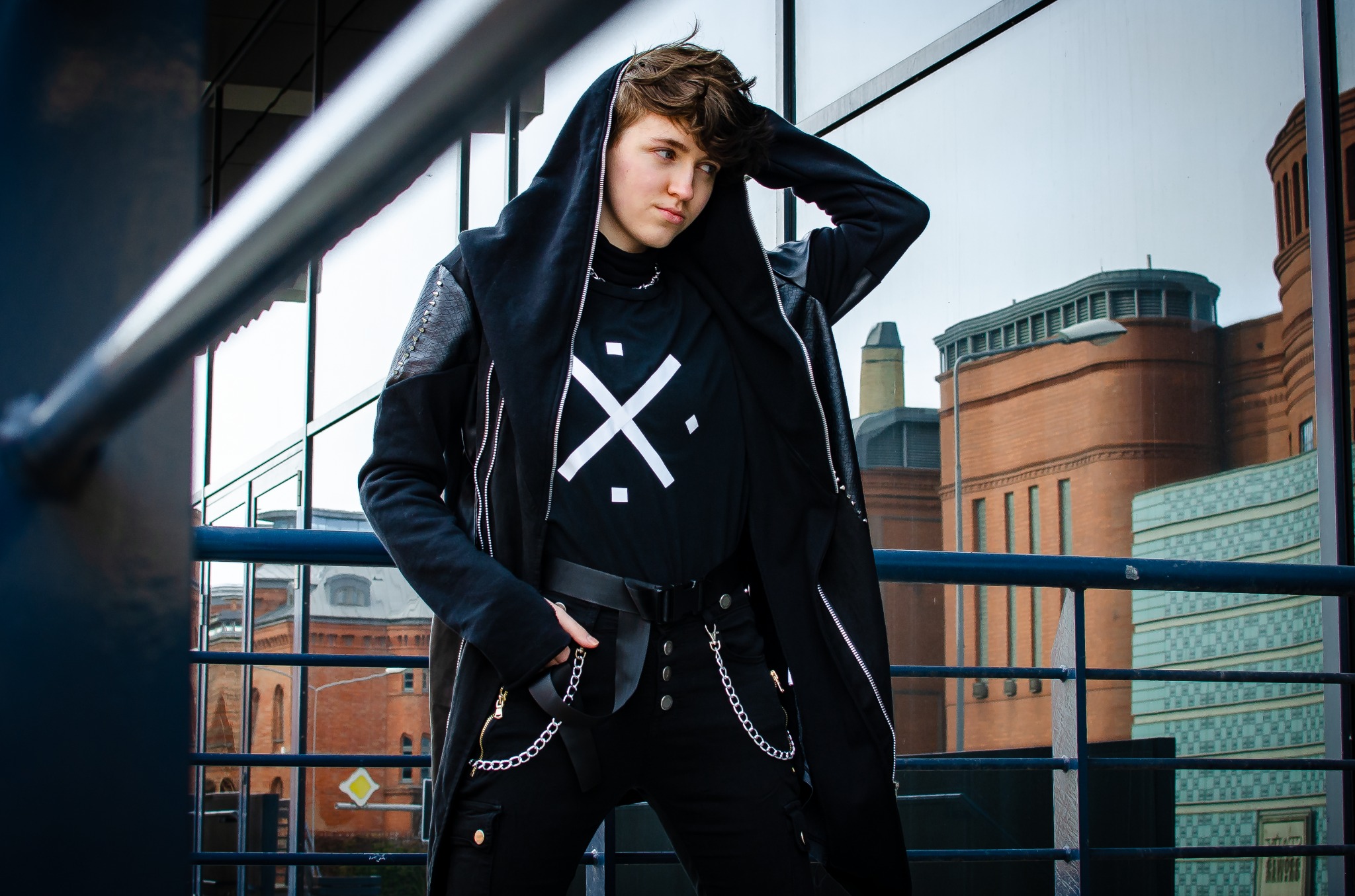 Osoba w czarnej, stylizowanej kurtce z kapturem i spodniach z łańcuchami, pozująca na tle miejskiej architektury i szklanych elewacji.