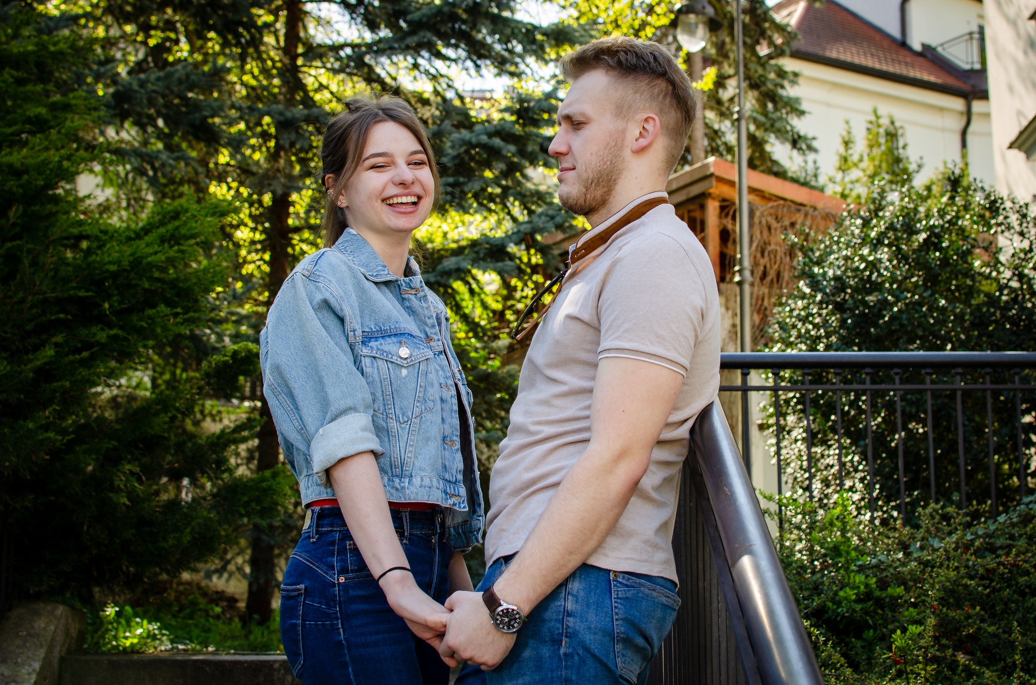 Uśmiechnięta młoda kobieta w dżinsowej kurtce trzyma za rękę mężczyznę opierającego się o metalową barierkę na tle zieleni i zabudowań.