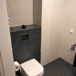 Nowoczesna toaleta z białym kompaktem WC, czarną zabudową geberit z chromowanym przyciskiem spłukującym i jasnoszarymi płytkami ściennymi z delikatnym wzorem.