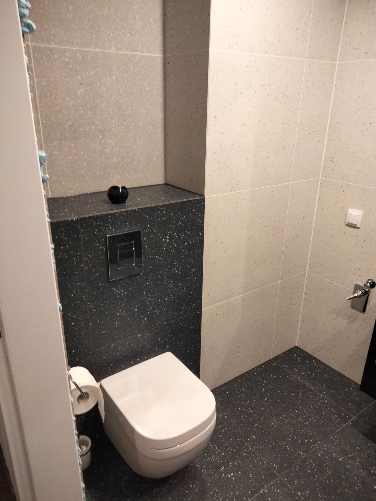 Nowoczesna toaleta z białym kompaktem WC, czarną zabudową geberit z chromowanym przyciskiem spłukującym i jasnoszarymi płytkami ściennymi z delikatnym wzorem.