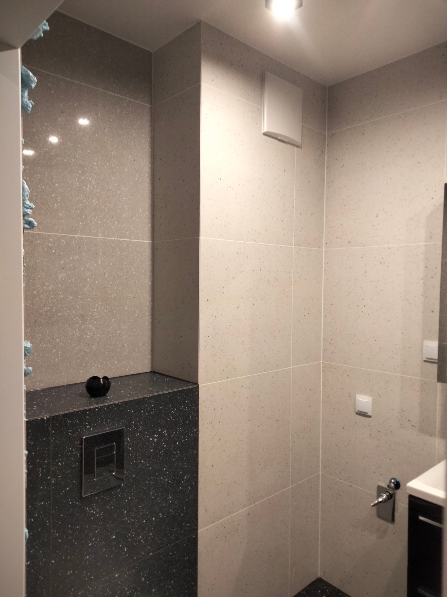 Nowoczesna łazienka z szarymi płytkami imitującymi kamień, czarnym blatem z ukrytym stelażem WC i minimalistyczną armaturą.