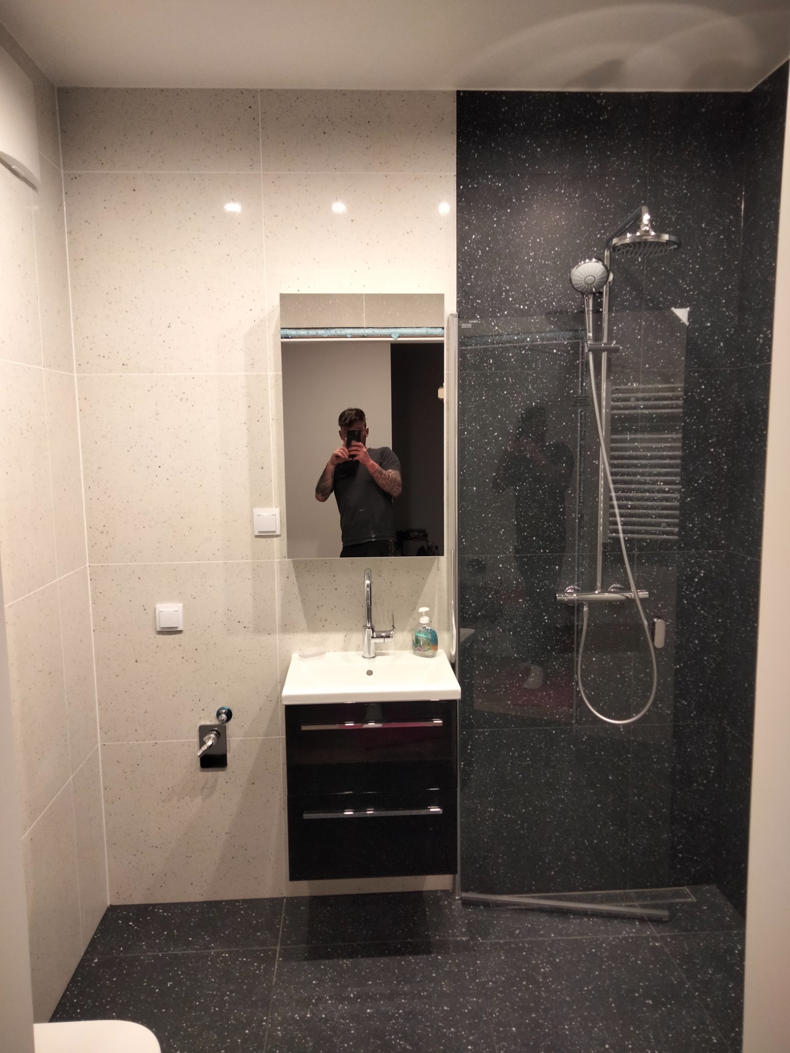 Nowoczesna łazienka z białymi i czarnymi płytkami imitującymi lastryko, kabina prysznicowa ze szklaną ścianką, umywalka z szafką, oraz odbicie fotografa w lustrze.