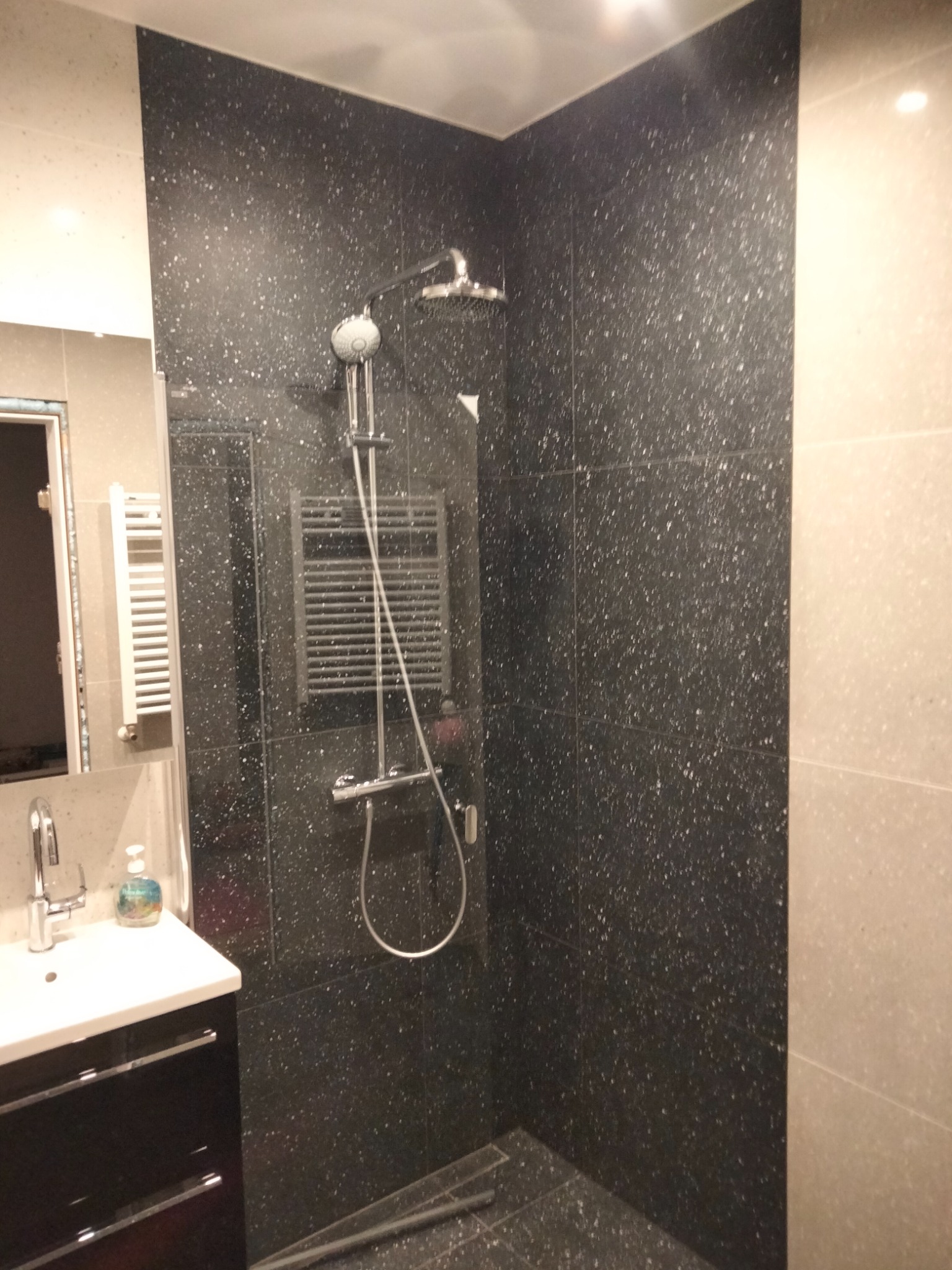 Nowoczesna kabina prysznicowa z czarnymi płytkami w drobne białe plamki i chromowaną armaturą, widoczna umywalka z szafką w Kościerzynie.