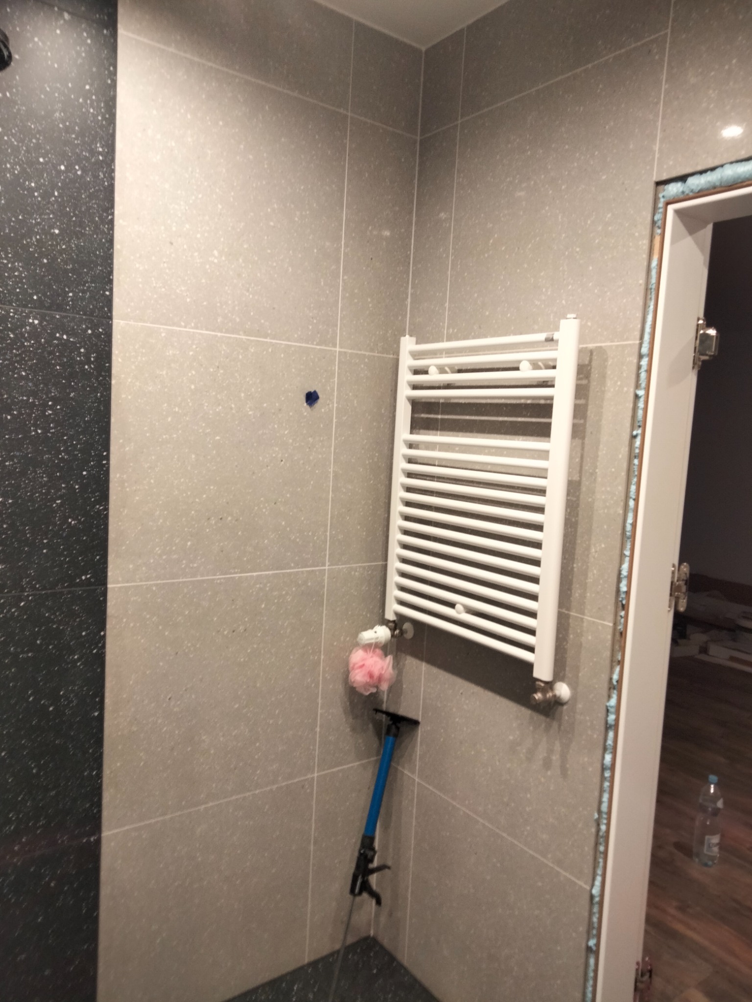 Wykończona łazienka z szarymi płytkami i czarnym akcentem, widoczny biały grzejnik drabinkowy, niebieski mop i różowa gąbka, drzwi z pianką montażową.