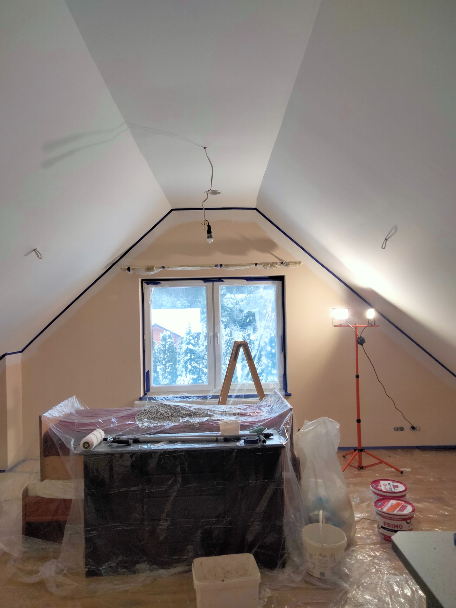 Poddasze zabezpieczone folią malarską, ściany częściowo pomalowane na beżowo, sufit na biało, widoczna drabina malarska, lampa halogenowa na statywie, puszki z farbą i narzędzia malarskie.