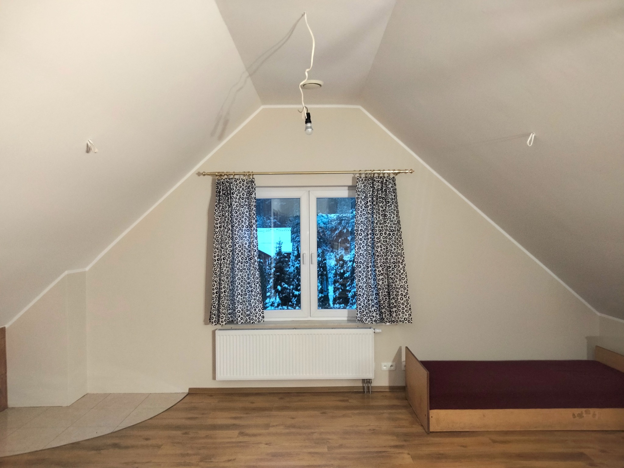 Poddasze po remoncie z oknem, kaloryferem i łóżkiem, widoczne niedoskonałości malowania na suficie i wiszący kabel od lampy.
