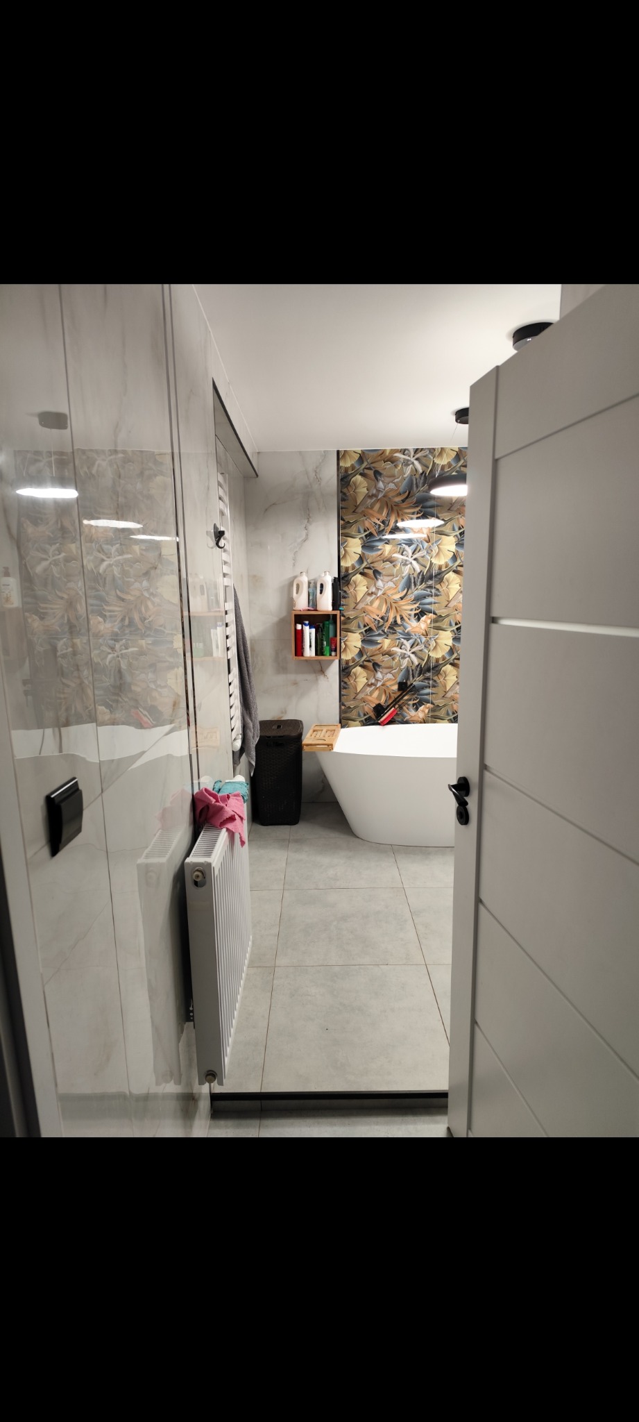 Nowoczesna łazienka z wanną wolnostojącą, marmurowymi płytkami i dekoracyjną ścianą w kwiatowe wzory. Widok zza uchylonych drzwi, grzejnik na ścianie.