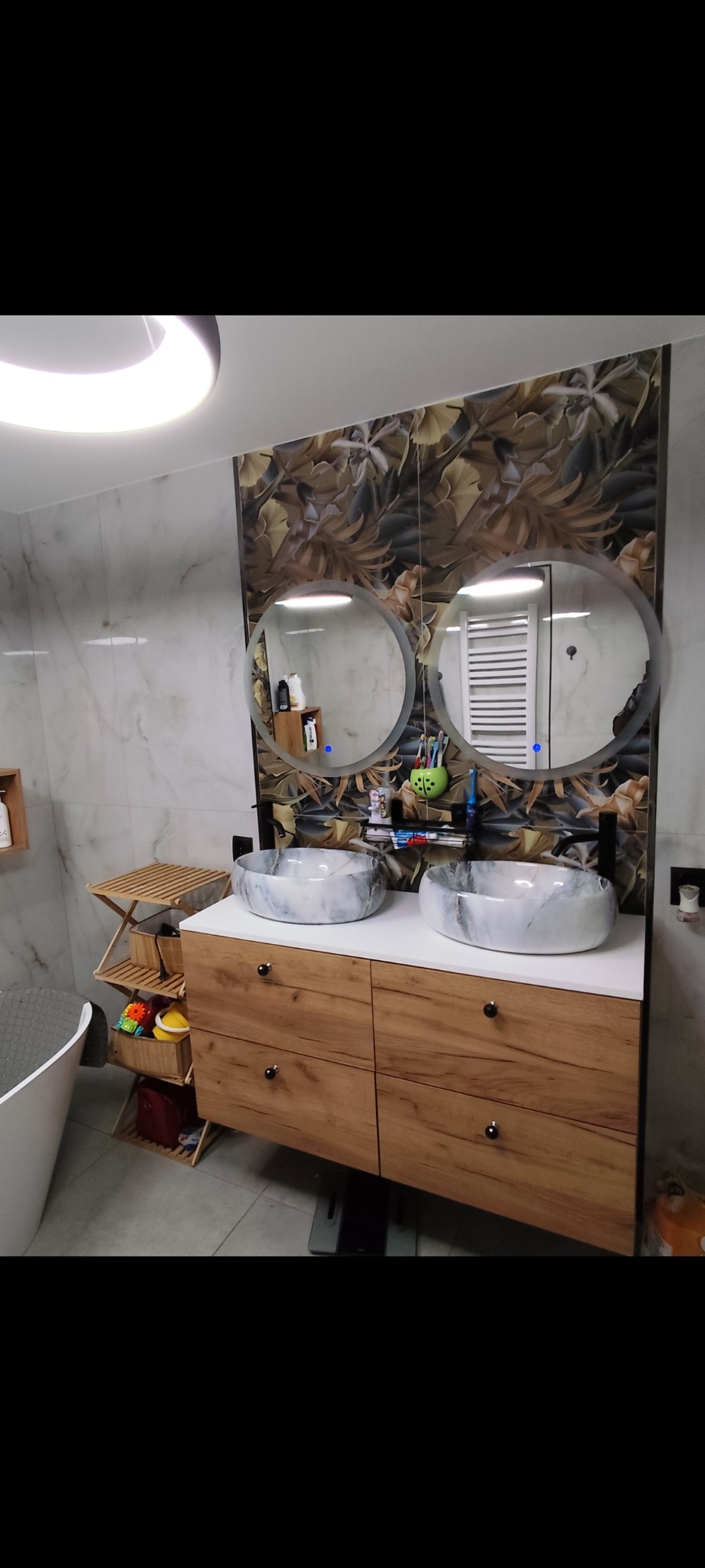 Wykończona łazienka z dwoma okrągłymi lustrami nad drewnianą szafką z umywalkami w stylu marmuru. Na ścianie płytki z motywem liści.